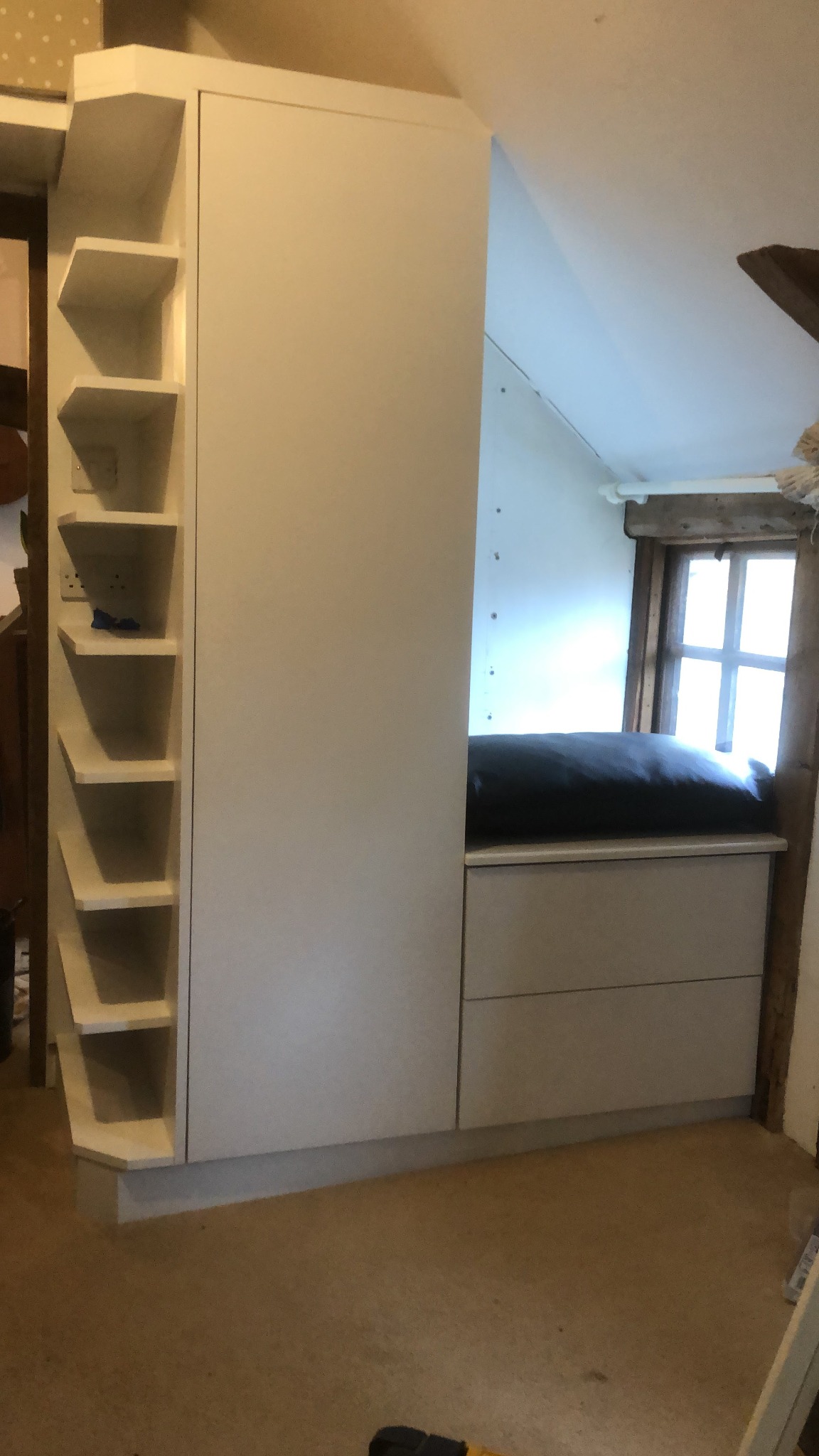 Biała zabudowa meblowa w skosie poddasza z otwartymi półkami i szafkami, widoczna instalacja elektryczna i okno z drewnianą ramą.