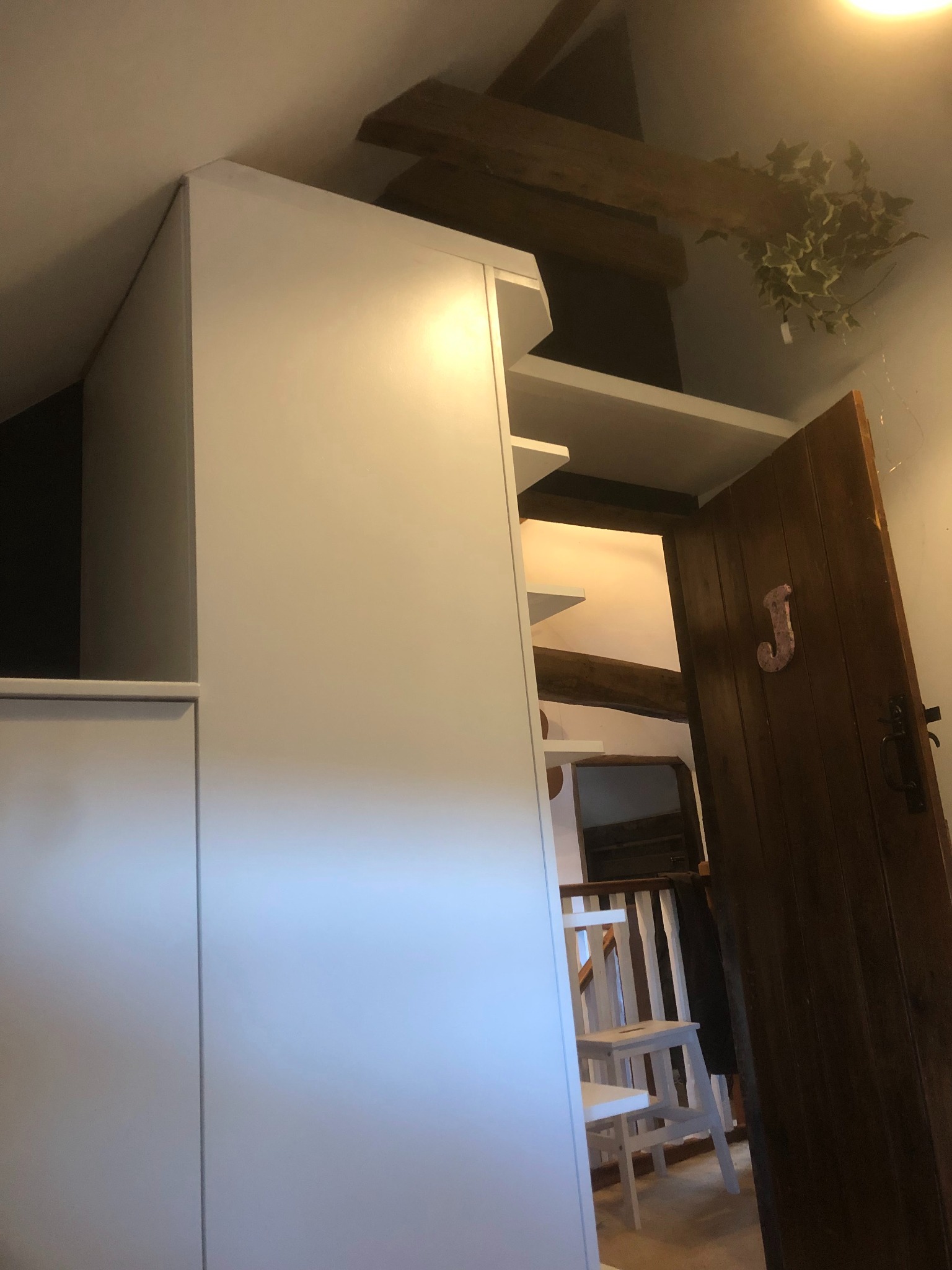 Biała szafa wnękowa z półkami, widoczna z perspektywy niskiego kąta, obok otwarta drewniana drzwi z ozdobną literą 'J', w tle widoczne drewniane belki stropowe i białe krzesło.