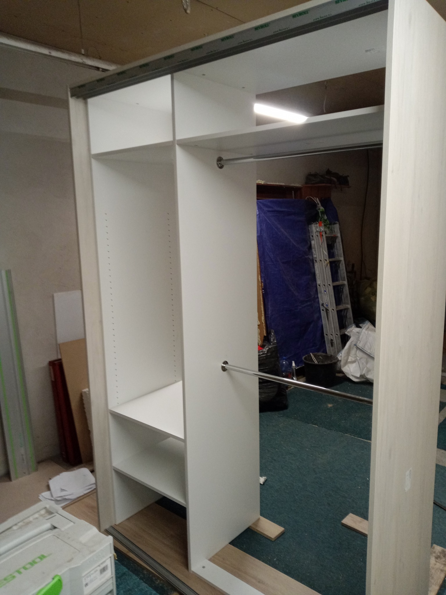 Montaż szkieletu szafy wnękowej z drążkami na ubrania, widoczne prowadnice przesuwne, w tle drabina i plandeka zabezpieczająca.