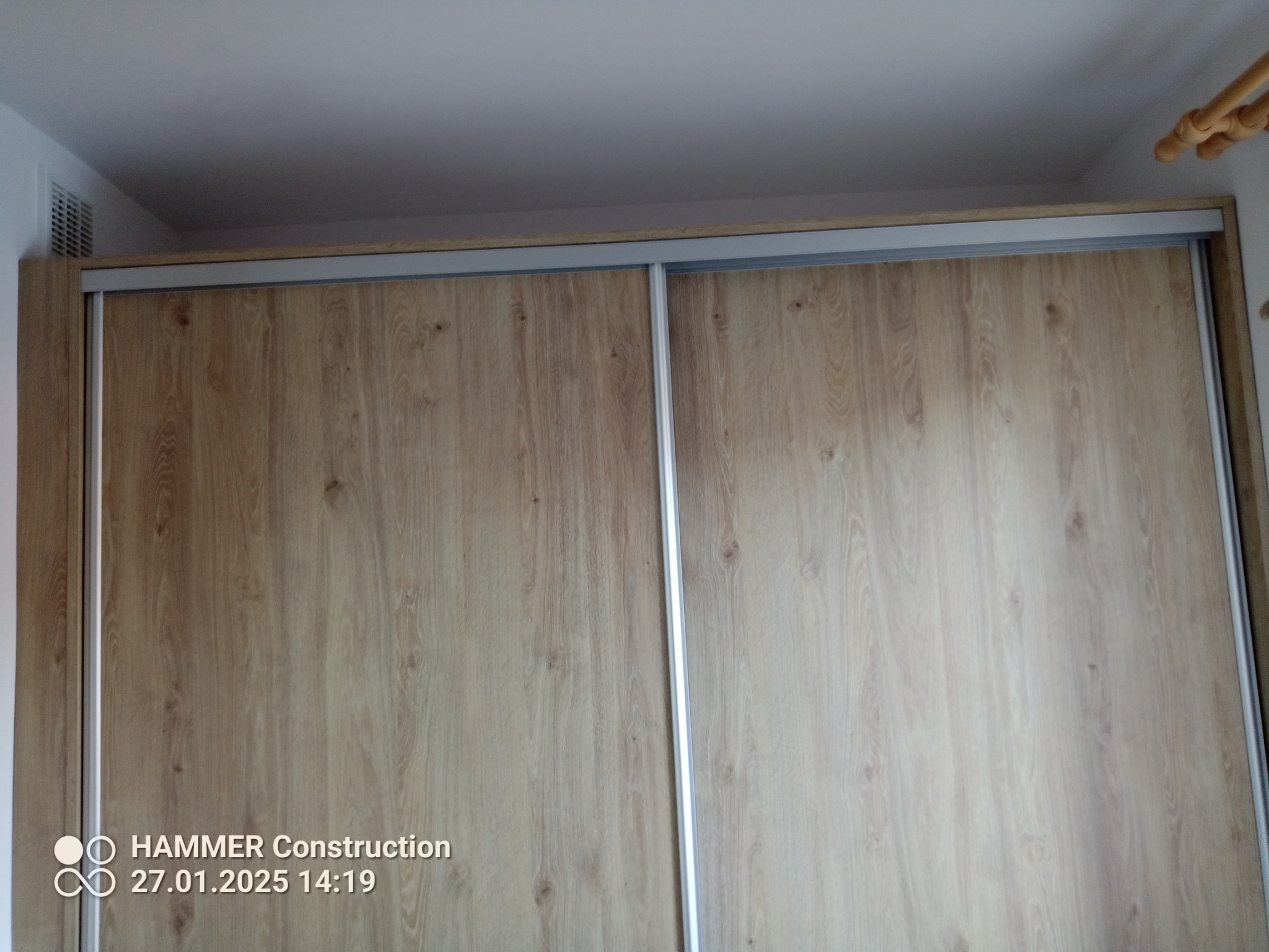 Zamknięta szafa wnękowa z przesuwnymi drzwiami w odcieniach jasnego drewna, widoczna kratka wentylacyjna na górze po lewej stronie.