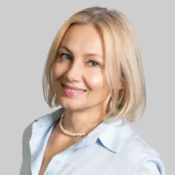 Portret uśmiechniętej, blondwłosej kobiety w błękitnej koszuli i perłowym naszyjniku na jednolitym, szarym tle.