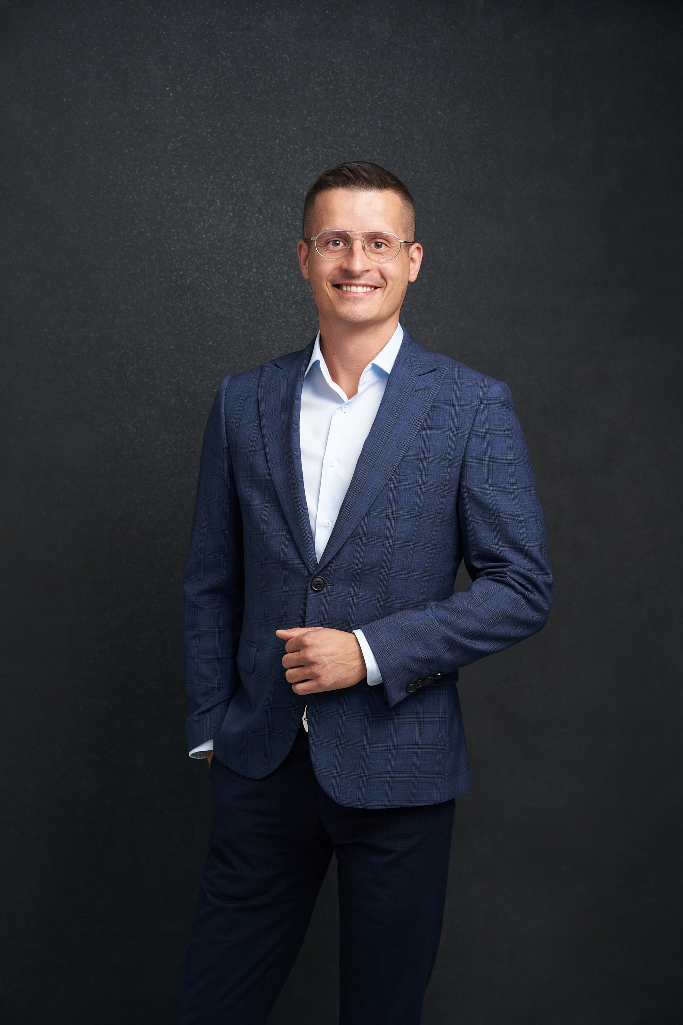 Uśmiechnięty mężczyzna w okularach i niebieskim garniturze w kratę, pozujący na ciemnoszarym tle.