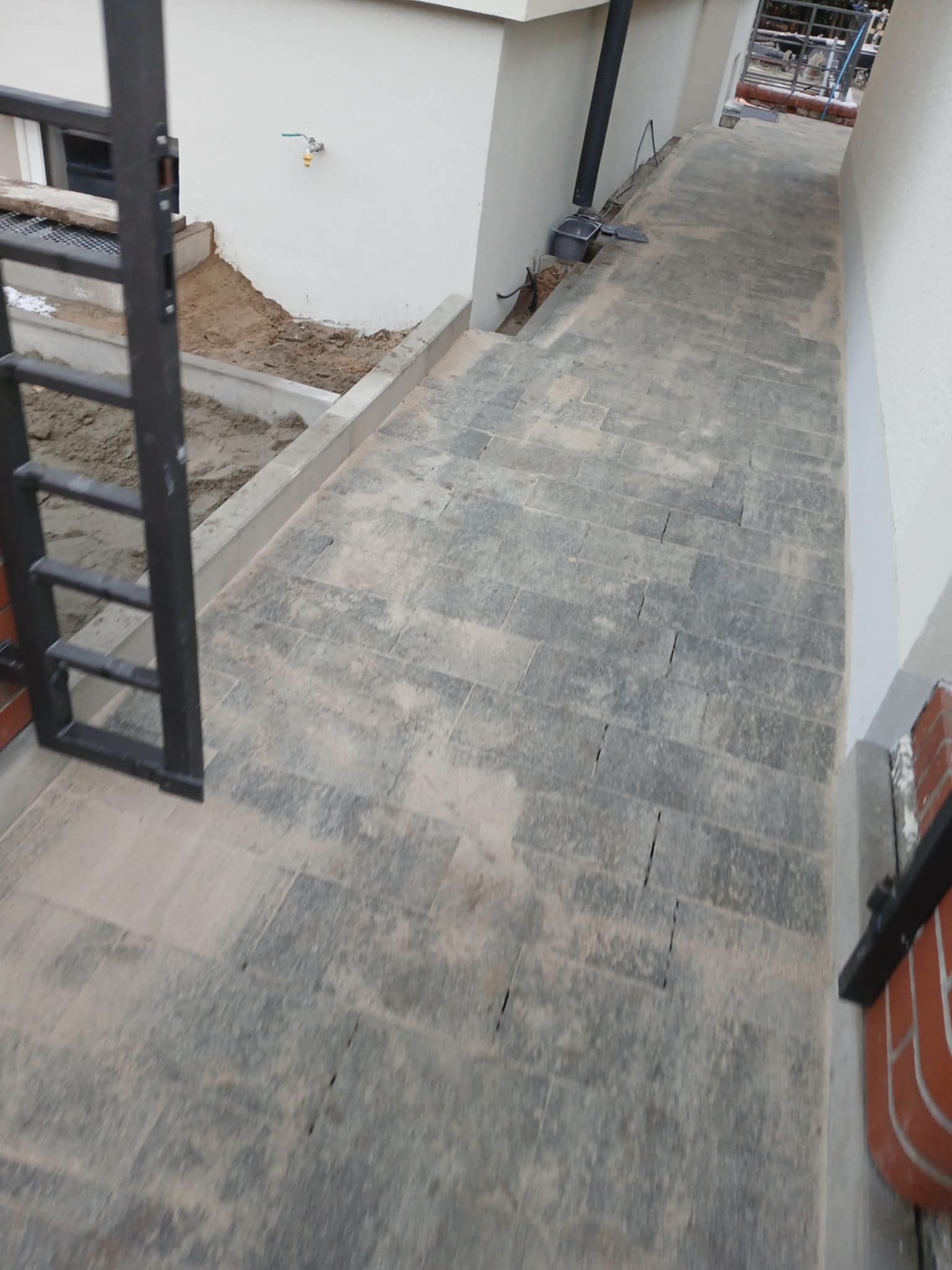 Ułożona szara kostka brukowa tworząca chodnik przy białym budynku, widoczna brama wejściowa z elementami piasku i instalacji.