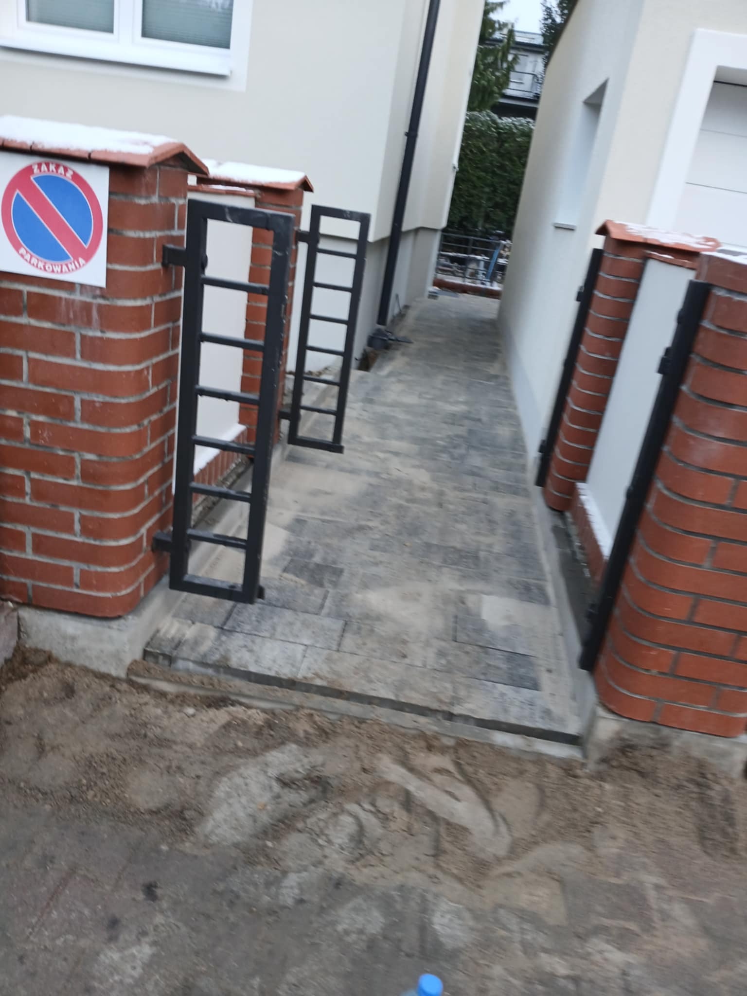 Ułożona szara kostka brukowa pomiędzy budynkami, z widocznym znakiem zakazu parkowania na ceglanym słupku i elementami furtki, teren wokół jeszcze niezagospodarowany.
