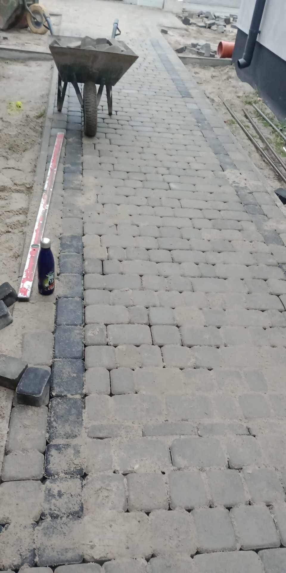 Świeżo ułożona ścieżka z kostki brukowej z ciemniejszym obramowaniem, taczka wypełniona kostką i poziomica obok, w tle fragment budynku.