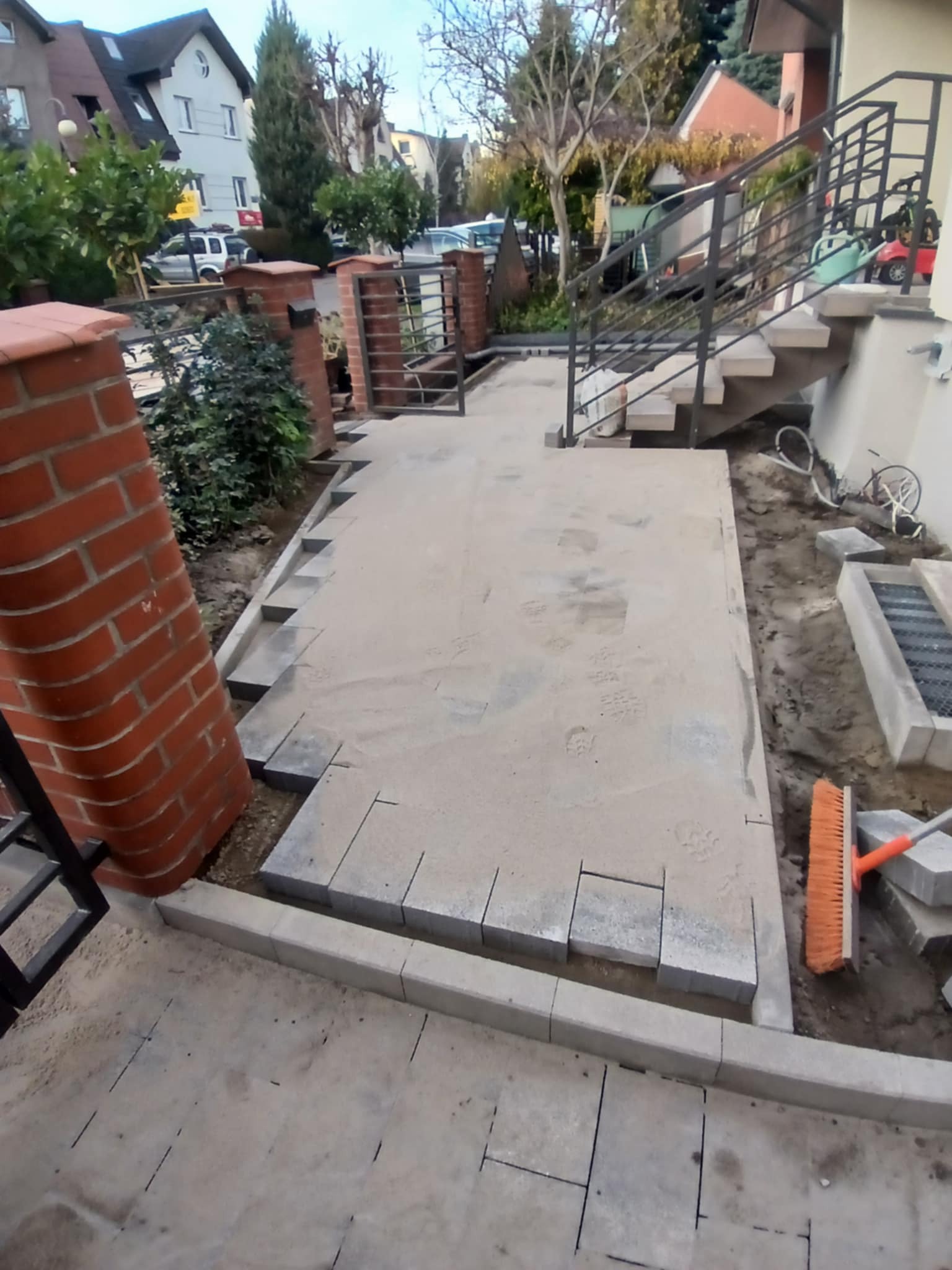 Przygotowany teren pod ułożenie kostki brukowej z widocznymi obrzeżami, piaskiem i częściowo ułożonymi kostkami, pomarańczowa zmiotka leży obok.