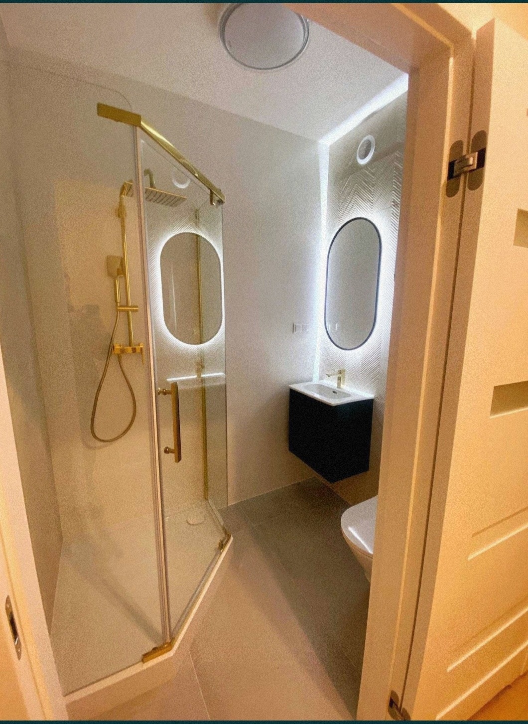 Nowoczesna łazienka z kabiną prysznicową ze złotymi detalami, podświetlanym lustrem i czarną szafką pod umywalką, widok zza uchylonych drzwi.