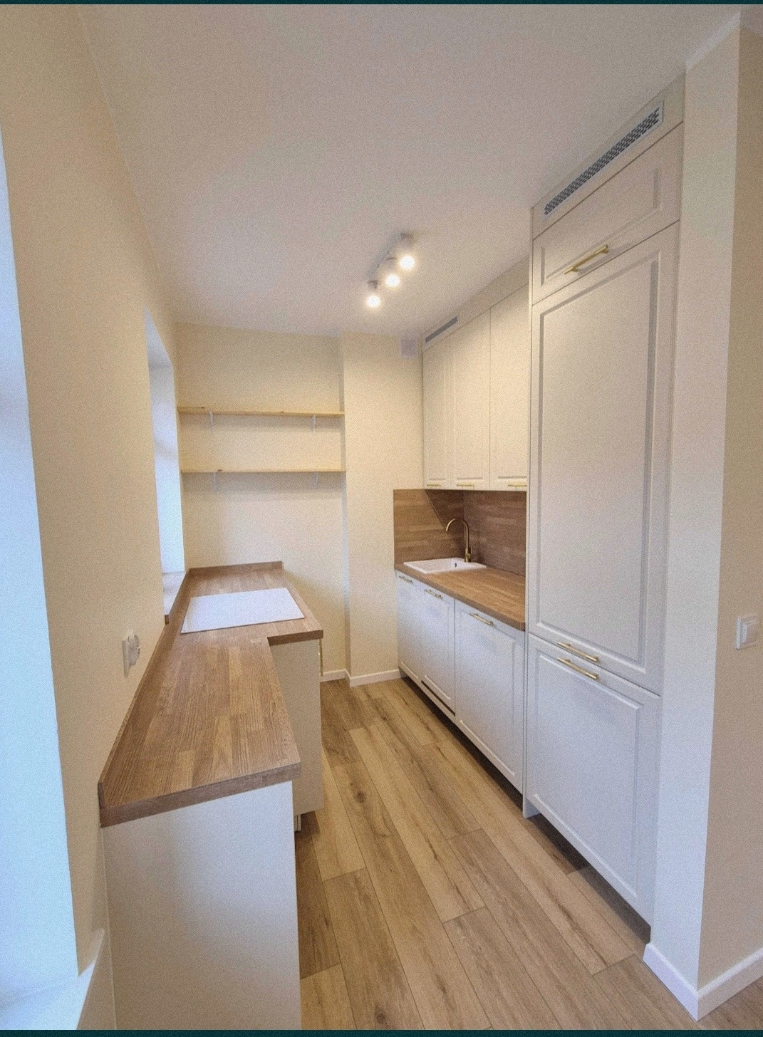 Jasna kuchnia z drewnianymi blatami i podłogą, białe szafki z złotymi uchwytami, półki wiszące na ścianie, oświetlenie punktowe.