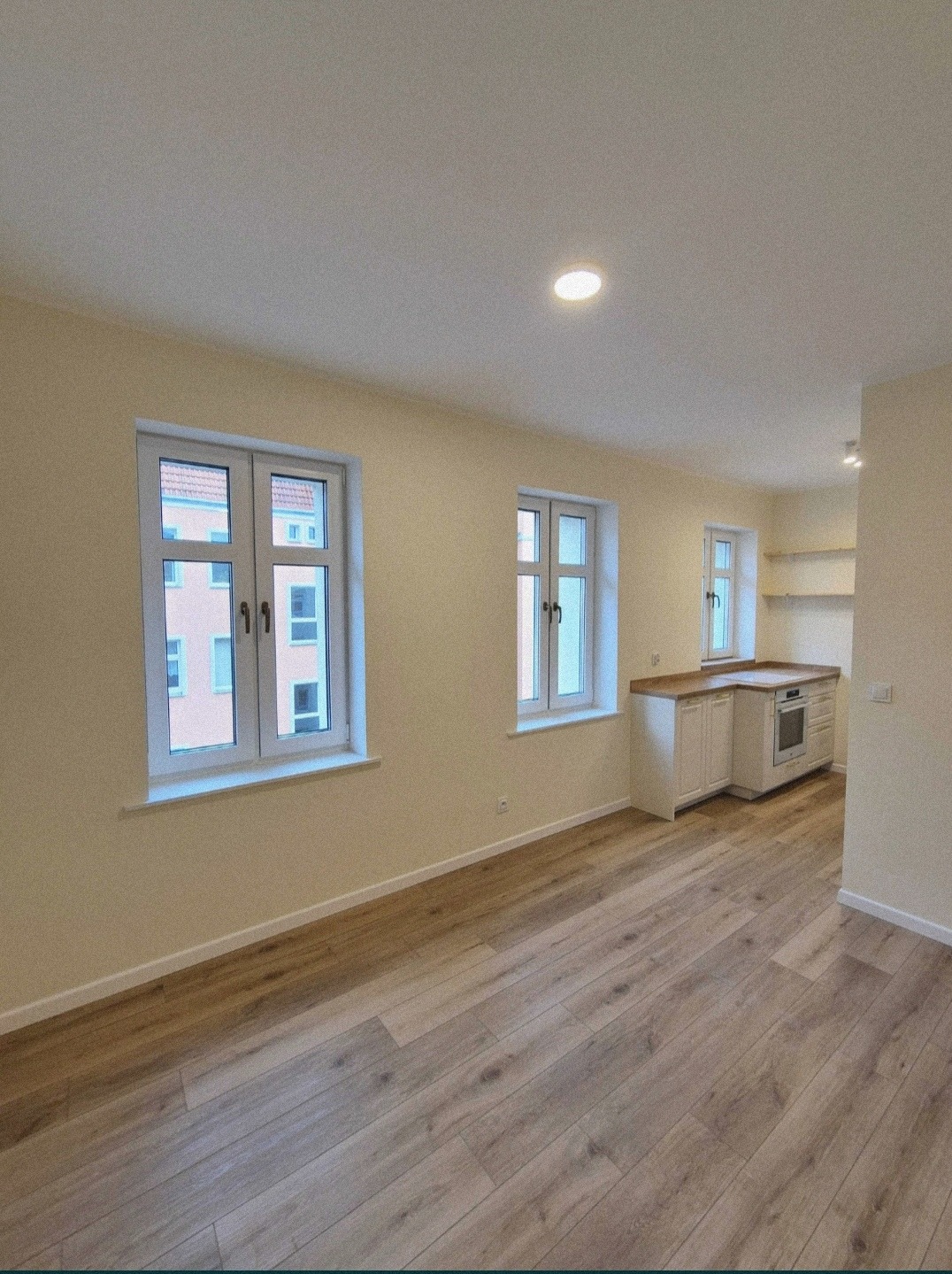 Jasne, przestronne wnętrze mieszkania po remoncie z drewnianą podłogą, trzema oknami z białymi ramami i widoczną zabudową kuchenną w głębi.