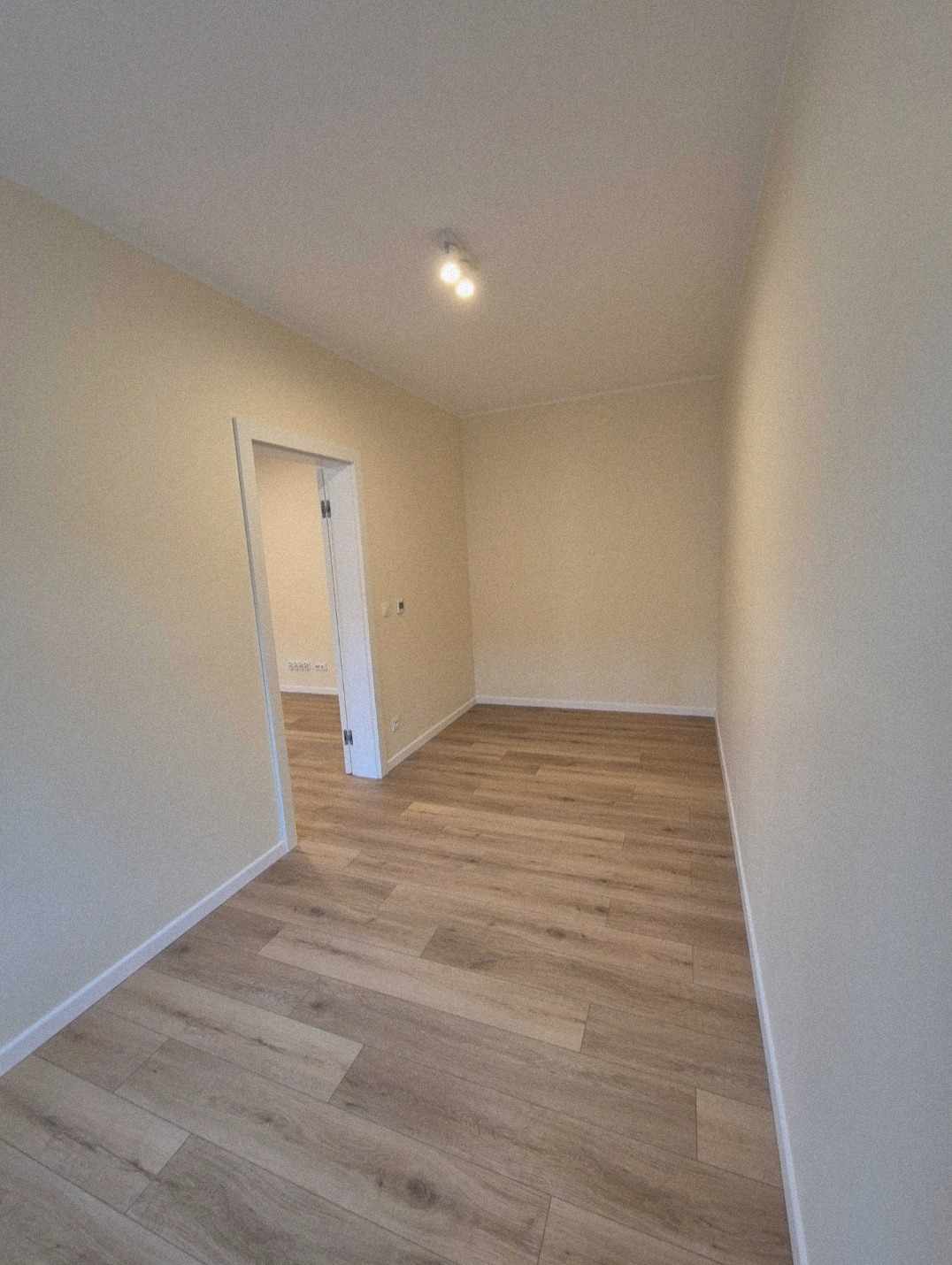 Pusty pokój po odświeżeniu z panelami podłogowymi imitującymi drewno ułożonymi wzdłuż, kremowe ściany i białe listwy przypodłogowe, widok na sąsiednie pomieszczenie przez otwarte drzwi.