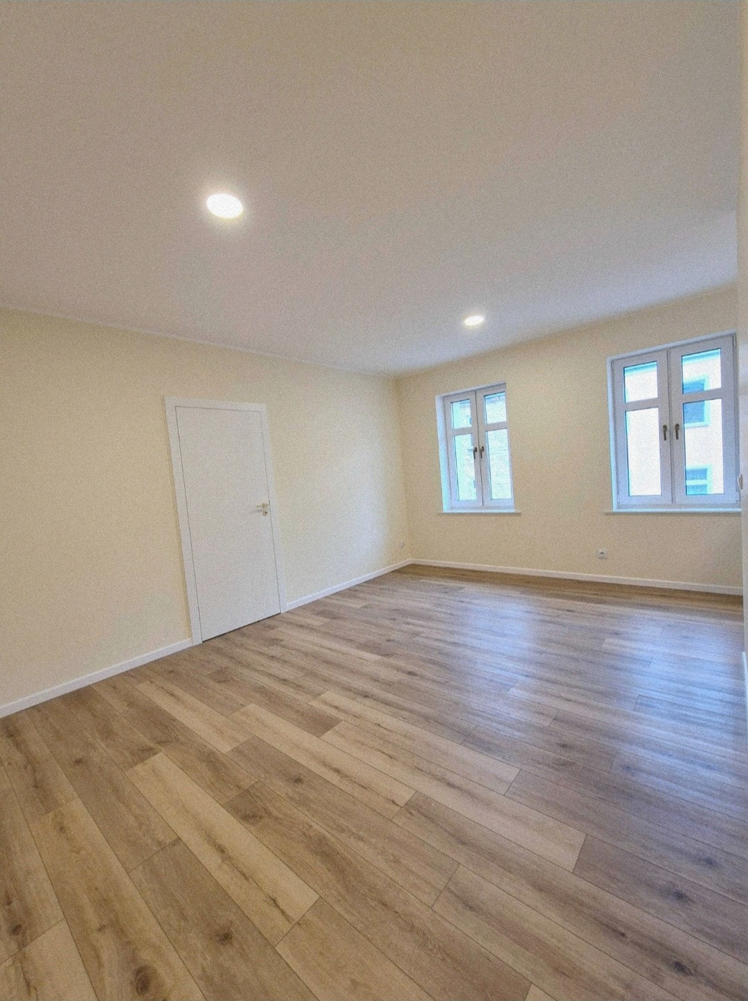 Pusty pokój po remoncie w Szczecinie z drewnopodobną podłogą, białymi ścianami i dwojgiem okien z widokiem na budynek.