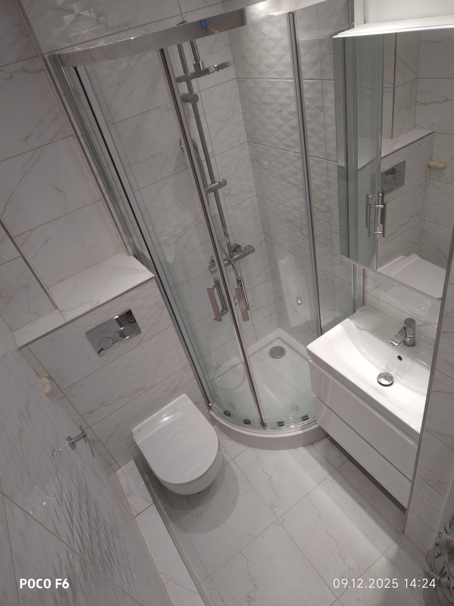 Nowoczesna, wykończona łazienka z kabiną prysznicową narożną, umywalką z szafką i podwieszaną toaletą. Białe marmurowe płytki na ścianach i podłodze.