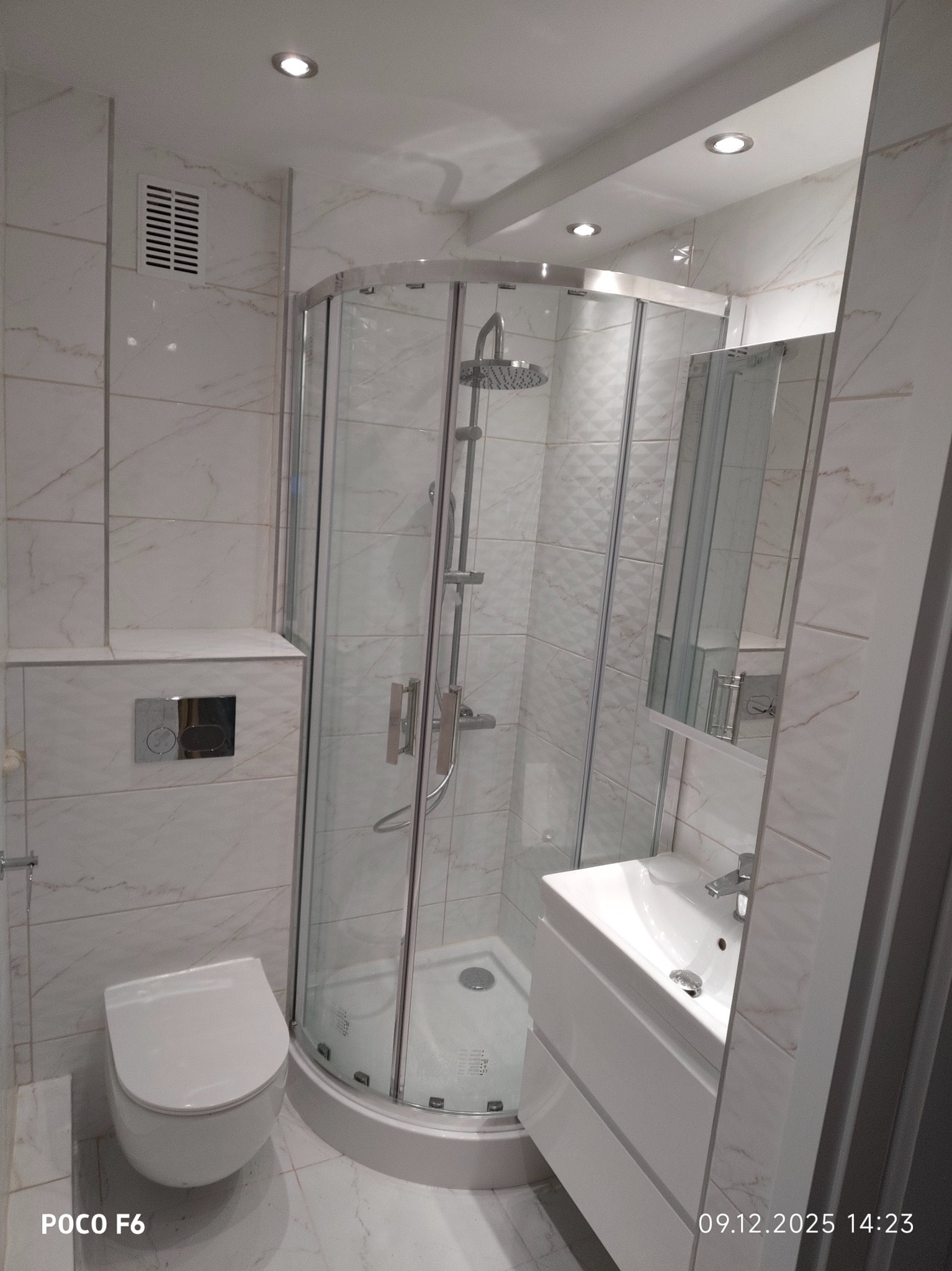 Nowoczesna łazienka po remoncie: kabina prysznicowa z hydromasażem, umywalka z szafką i podwieszana toaleta. Białe marmurowe płytki na ścianach i podłodze.