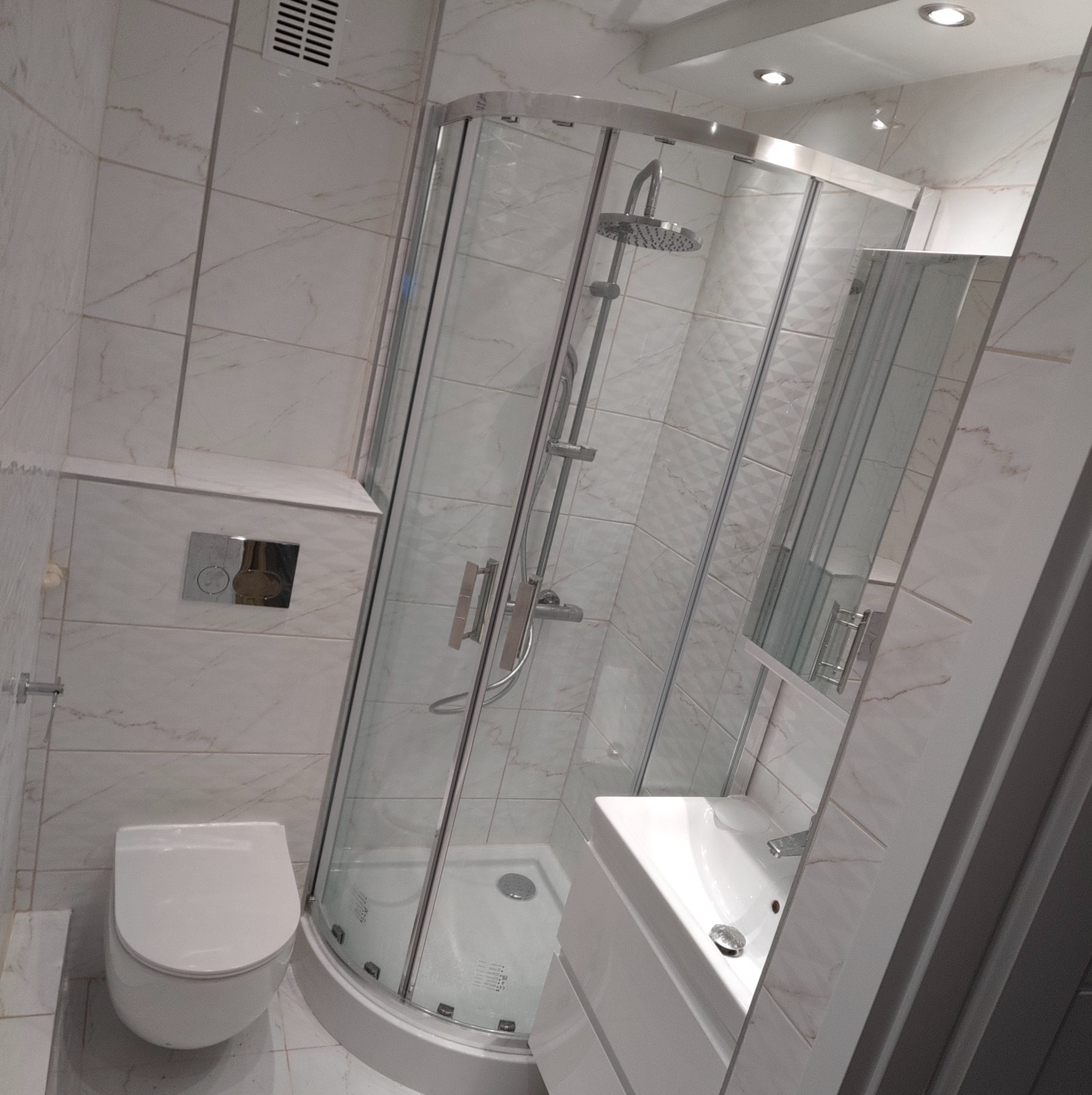 Jasna łazienka po remoncie: kabina prysznicowa z deszczownicą, podwieszana toaleta i umywalka z szafką. Białe płytki z delikatnym wzorem marmuru.