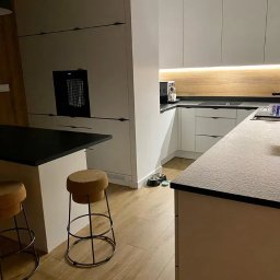 POLCRAFT - Remonty i wykończenia wnętrz - Nowoczesna kuchnia z białymi szafkami, czarnym blatem i drewnianymi akcentami, oświetlona listwą LED pod szafkami górnymi, z wyspą i hokerami oraz miskami dla zwierząt w rogu.