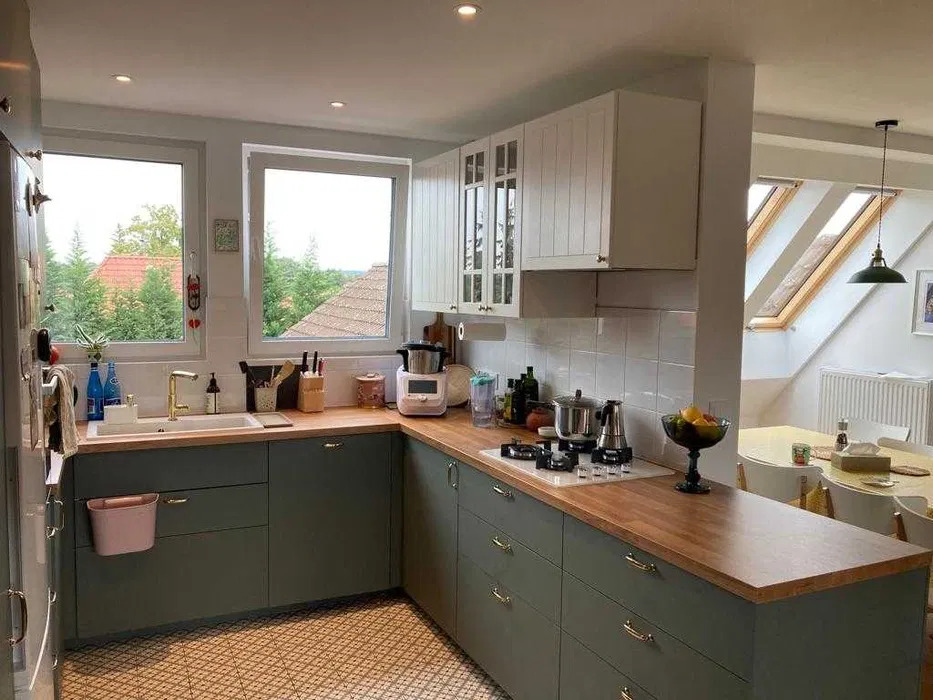 Nowoczesna kuchnia w odcieniach zieleni i bieli z drewnianym blatem, widok na ogród przez okno, na blacie kuchenka gazowa i naczynia, podłoga wyłożona wzorzystymi płytkami.