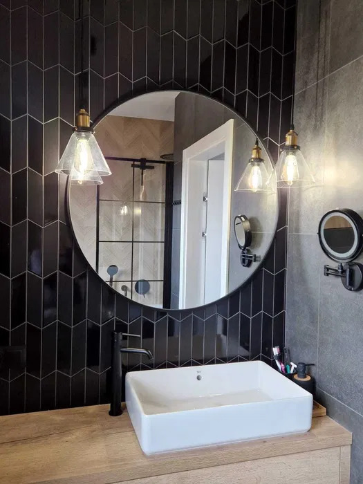 Nowoczesna łazienka w Szczecinie z czarnymi płytkami ułożonymi w jodełkę, okrągłym lustrem i wiszącymi lampami z przezroczystymi kloszami, białą prostokątną umywalką nablatową i czarną armaturą.