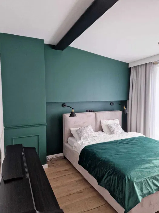 Sypialnia z zielonymi ścianami, drewnianą podłogą i łóżkiem z welurowym zagłówkiem, oświetlona czarnymi kinkietami