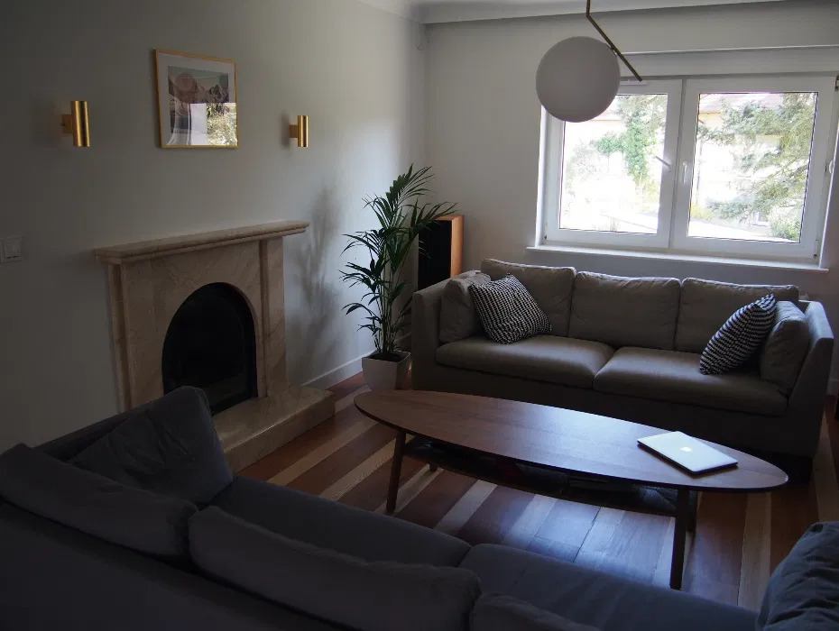 Jasne wnętrze salonu z kominkiem obłożonym beżowym marmurem, szarą sofą, drewnianym stolikiem kawowym z laptopem i parkietem w odcieniach brązu.