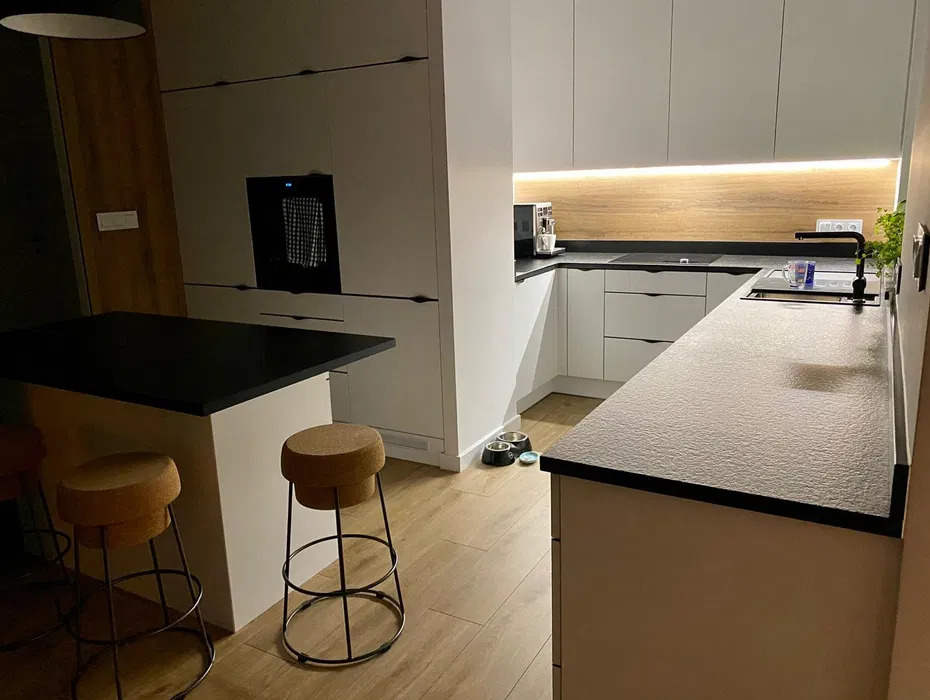 Nowoczesna kuchnia z białymi szafkami, czarnym blatem i drewnianymi akcentami, oświetlona listwą LED pod szafkami górnymi, z wyspą i hokerami oraz miskami dla zwierząt w rogu.