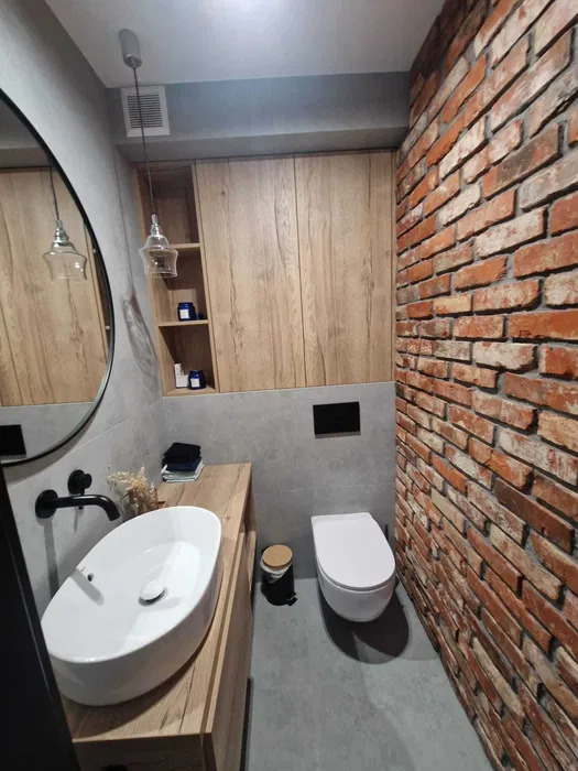 Nowoczesna łazienka z umywalką nablatową, drewnianą szafką, toaletą podwieszaną i ścianą z czerwonej cegły, oświetlona wiszącymi lampami z przezroczystego szkła.