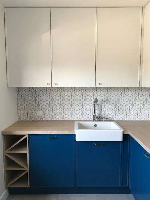 Nowoczesna łazienka z białymi szafkami górnymi, granatowymi szafkami dolnymi z drewnianym blatem i białą ceramiczną umywalką farmerską, geometryczne biało-szare płytki na ścianie.