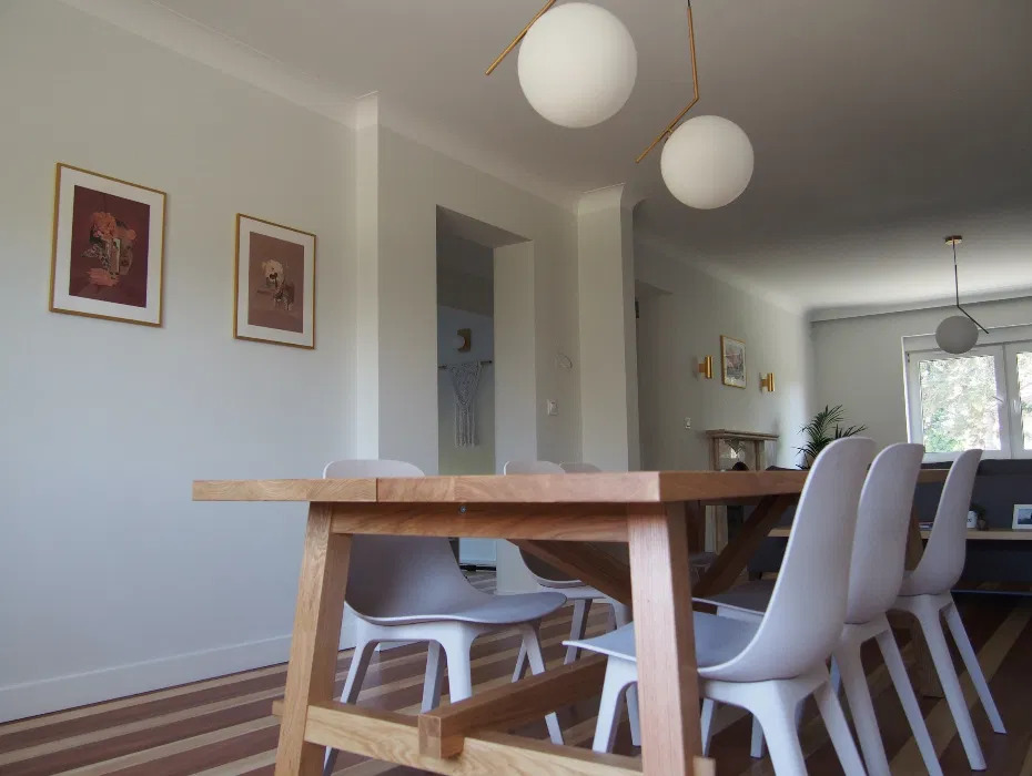 Jasne wnętrze jadalni z drewnianym stołem i białymi krzesłami, parkiet w paski, na ścianie dwa obrazy w złotych ramach, kuliste lampy sufitowe.