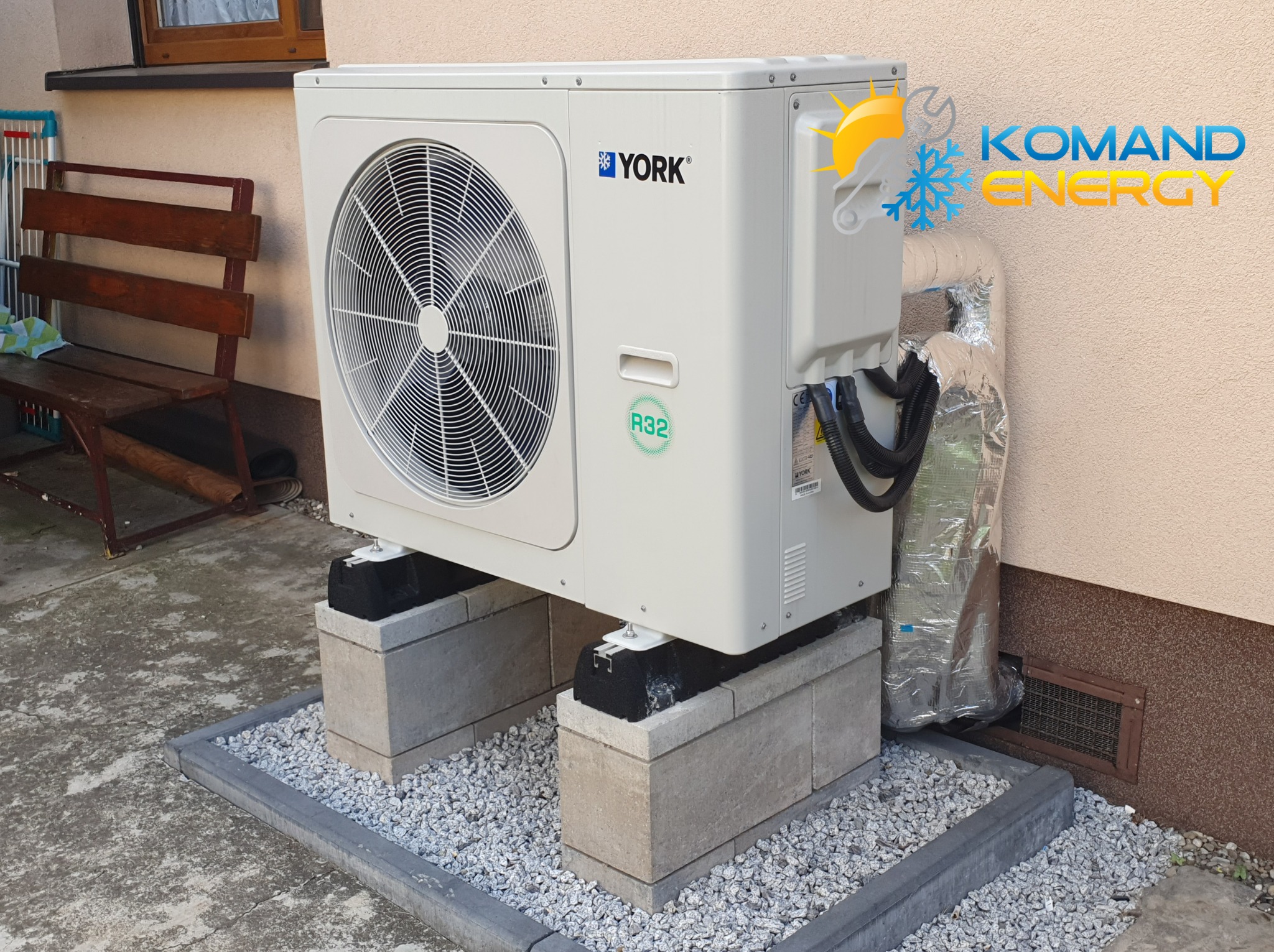 Biała pompa ciepła YORK R32 na betonowych blokach, z logo firmy Komand Energy w rogu, na tle beżowej ściany budynku i szarego chodnika z kamykami.
