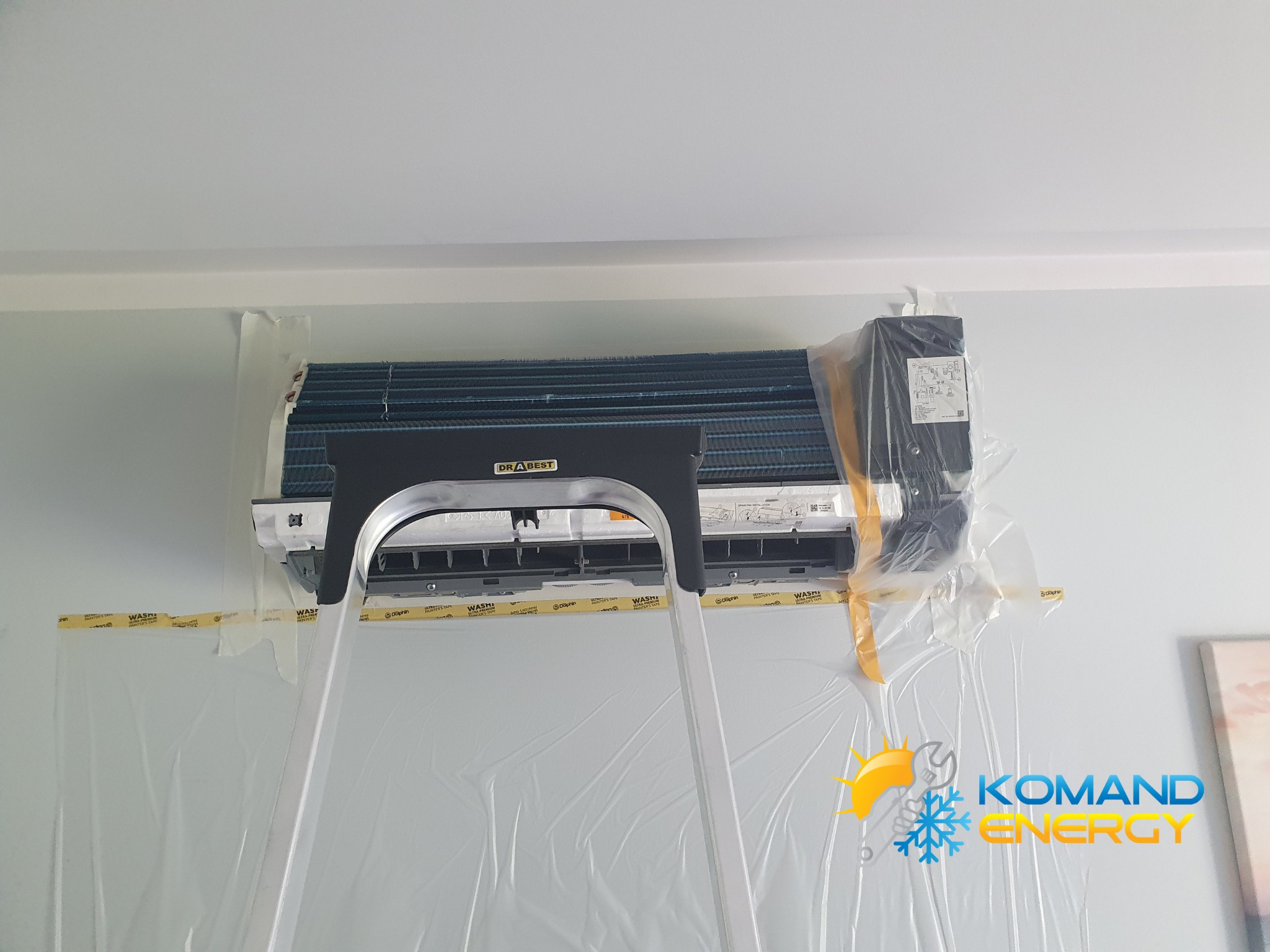 Klimatyzator ścienny marki DrA Best, zabezpieczony folią i taśmą malarską, z drabiny podczas montażu. Widoczne detale instalacji i logo firmy na ścianie.