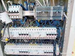 Rozdzielnica elektryczna z licznymi bezpiecznikami i okablowaniem, widoczne oznaczenia ostrzegawcze na obudowach, ujęcie z bliska.