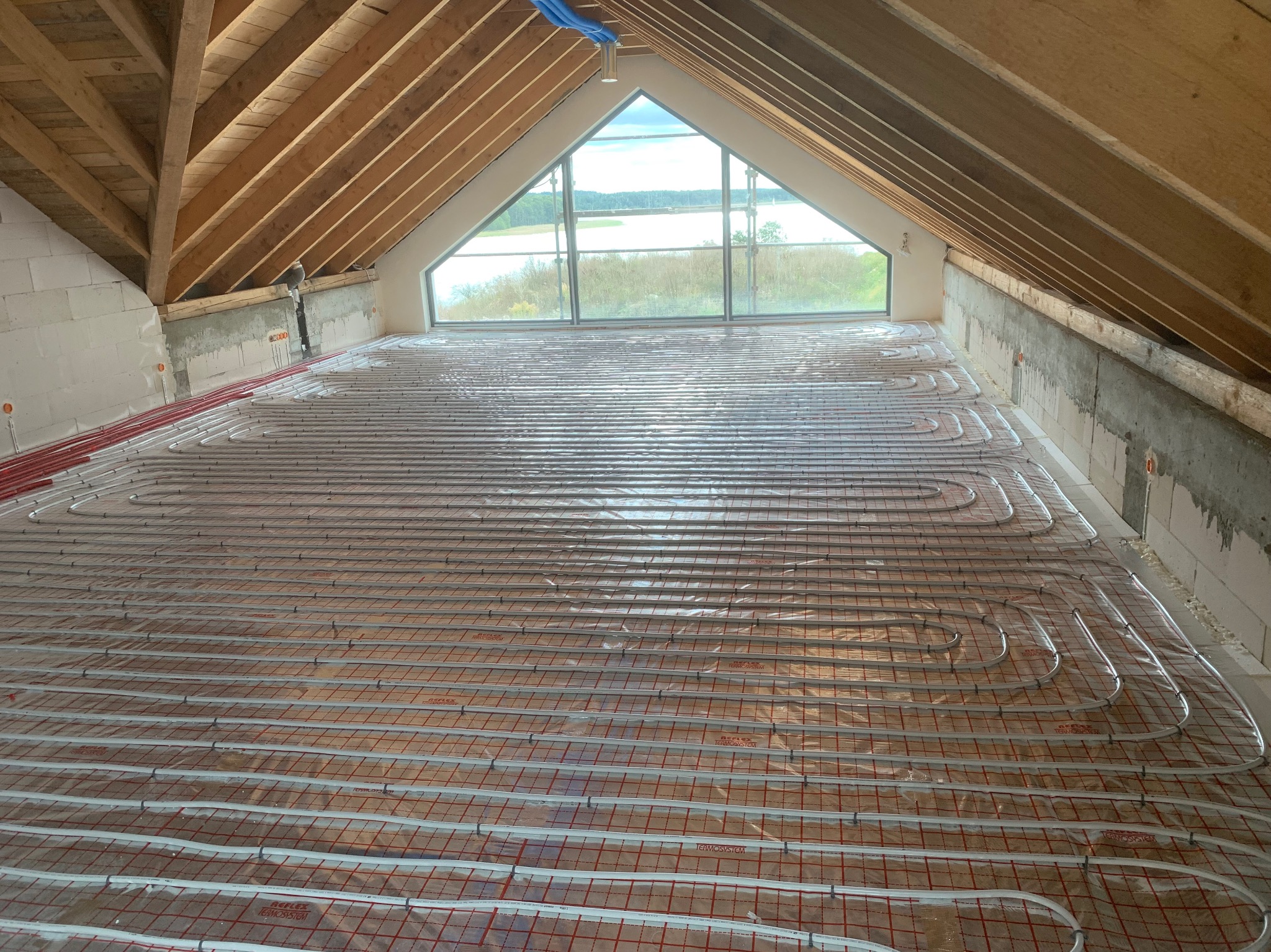 Montaż ogrzewania podłogowego na poddaszu z widokiem na jezioro przez trójkątne okno, rury grzewcze ułożone na folii z czerwoną siatką, surowy stan ścian z betonu komórkowego, widoczne krokwie dachowe