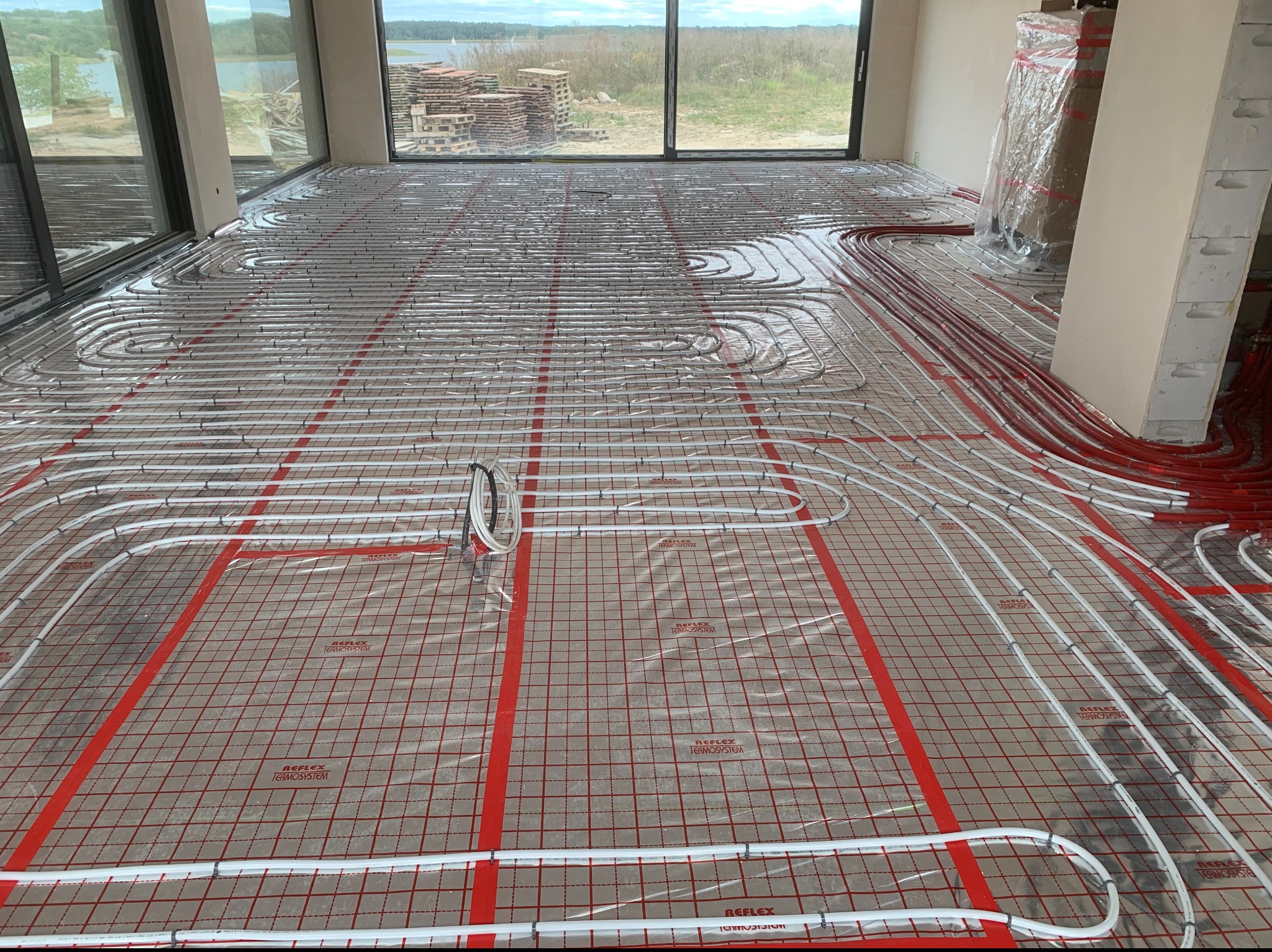 Rozłożone rury ogrzewania podłogowego na folii z czerwoną kratką w surowym wnętrzu z dużymi oknami i widokiem na jezioro oraz stertę palet.