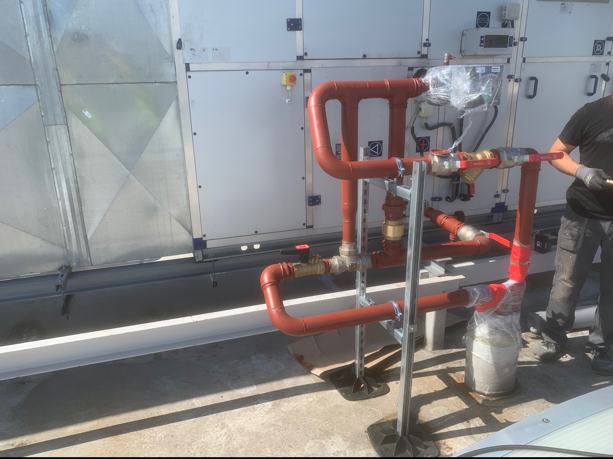 Instalacja rurociągów z zaworami, częściowo owinięta folią ochronną, na tle centrali wentylacyjnej z pracownikiem w rękawicach, widok z góry na płaskim dachu.