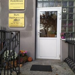 Kancelaria Doradztwa Podatkowego DOMBROWSKA&WOLFRAM SC - Wejście do biura rachunkowego i doradcy podatkowego z tabliczkami informacyjnymi, kwiatami w donicach i dynią dekoracyjną przed drzwiami.