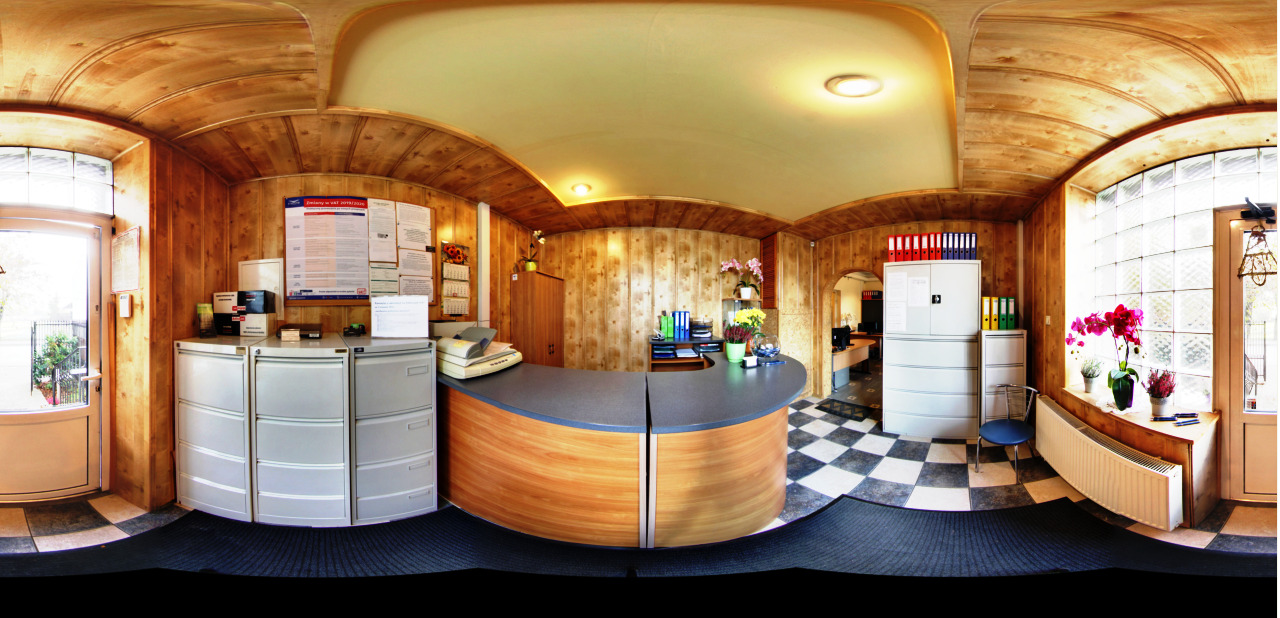 Panoramiczne ujęcie biura z drewnianą boazerią, recepcją z szarą ladą i drewnianym frontem, metalowymi szafkami na dokumenty, kwiatami w doniczkach na parapecie i czarno-białą podłogą.