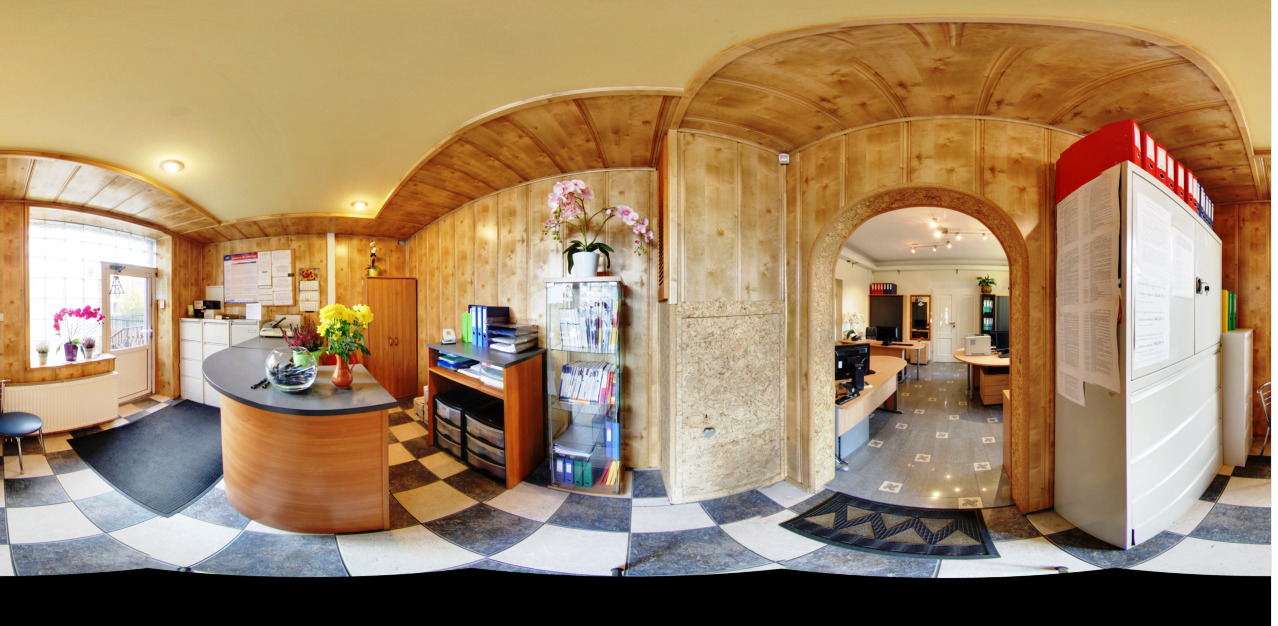 Panoramiczny widok wnętrza biura z drewnianą boazerią, recepcją z kwiatami, regałami z dokumentami i biurkami w dalszej części pomieszczenia.