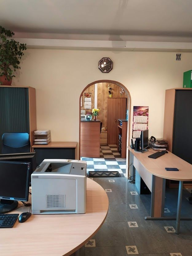 Biuro z dwoma biurkami, komputerami, drukarką, szafami na dokumenty, zegarem na ścianie i rośliną doniczkową w rogu, z widokiem na dalszą część biura przez łukowate przejście.