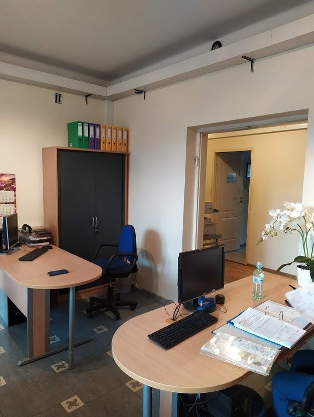 Biuro z dwoma biurkami, komputerami, segregatorami i kwiatami, widok na drzwi do innego pomieszczenia.