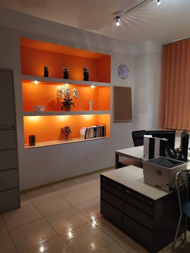 Nowoczesne biuro rachunkowe z pomarańczowymi wnękami na ścianie, w których ustawiono dekoracje, kwiaty i książki, biurka z komputerami i drukarką.