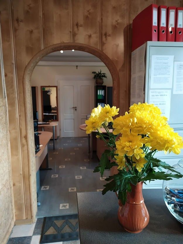 Wazon z żółtymi chryzantemami na biurku w biurze z widocznymi segregatorami i dokumentami, z perspektywą przez łukowe przejście do dalszej części pomieszczenia.