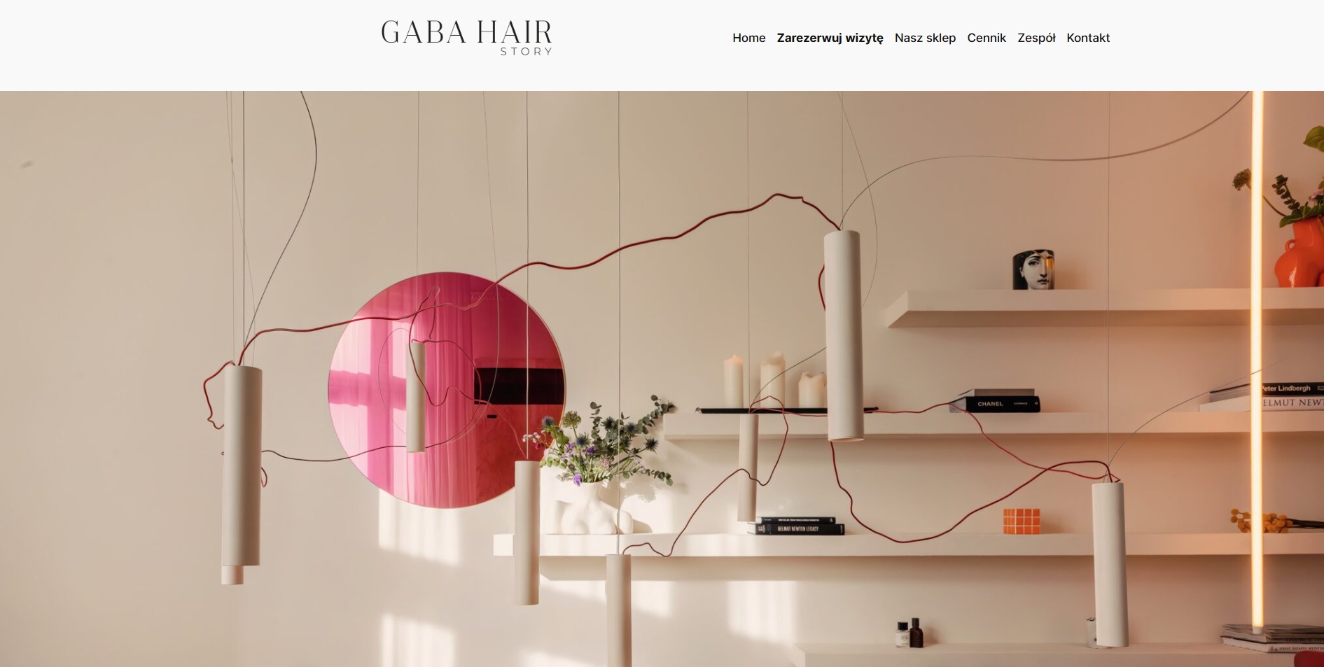 Nowoczesne wnętrze salonu fryzjerskiego z minimalistycznym designem, wiszące cylindryczne lampy z czerwonymi kablami, półki z książkami i dekoracjami, okrągłe różowe lustro.