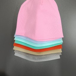 Szpula u Danusi Danuta Starkbauer - Stos czapek z dzianiny prążkowanej w pastelowych kolorach (różowy, błękitny, miętowy, pomarańczowy, beżowy, szary) na szarym tle. Widok z góry, wyraźna faktura materiału.