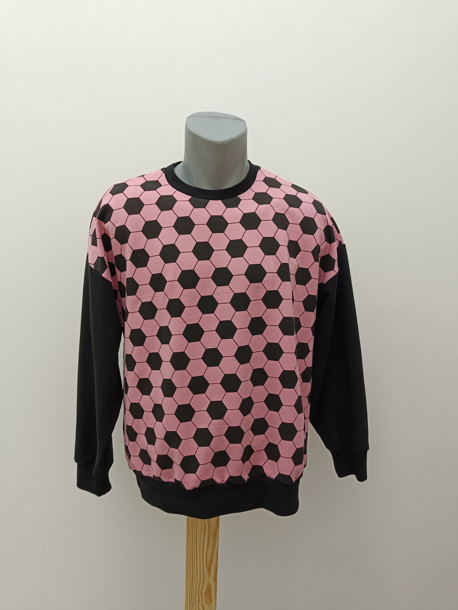 Bluza z długim, czarnym rękawem i wzorem różowo-czarnych heksagonów, prezentowana na manekinie na tle białej ściany. Wyeksponowany krój i deseń tkaniny.