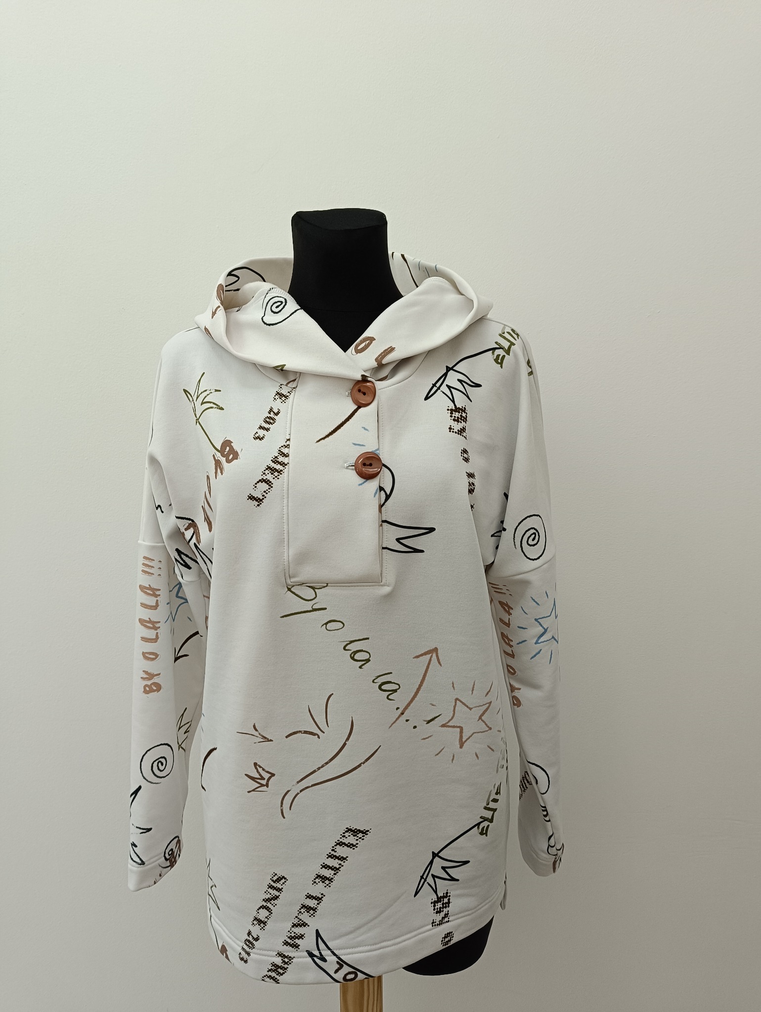 Biała bluza z kapturem, z brązowymi guzikami i kolorowymi nadrukami, prezentowana na czarnym manekinie na tle jasnej ściany. Widoczne napisy i graficzne elementy.