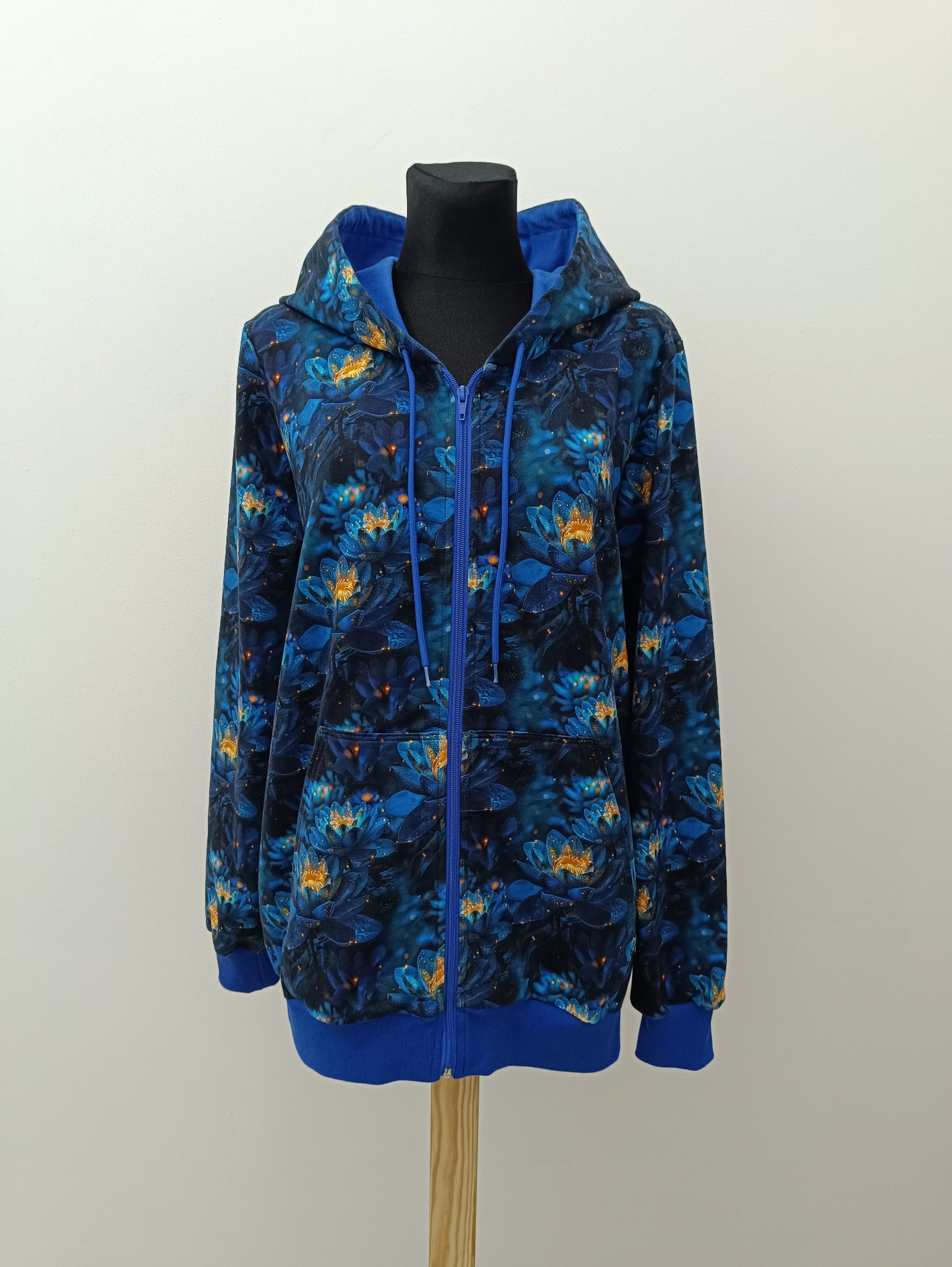 Rozpięta, niebieska bluza z kapturem, z motywem kwiatowym na czarnym manekinie. Bluza ma kieszenie i jest zaprezentowana na drewnianej podstawie.