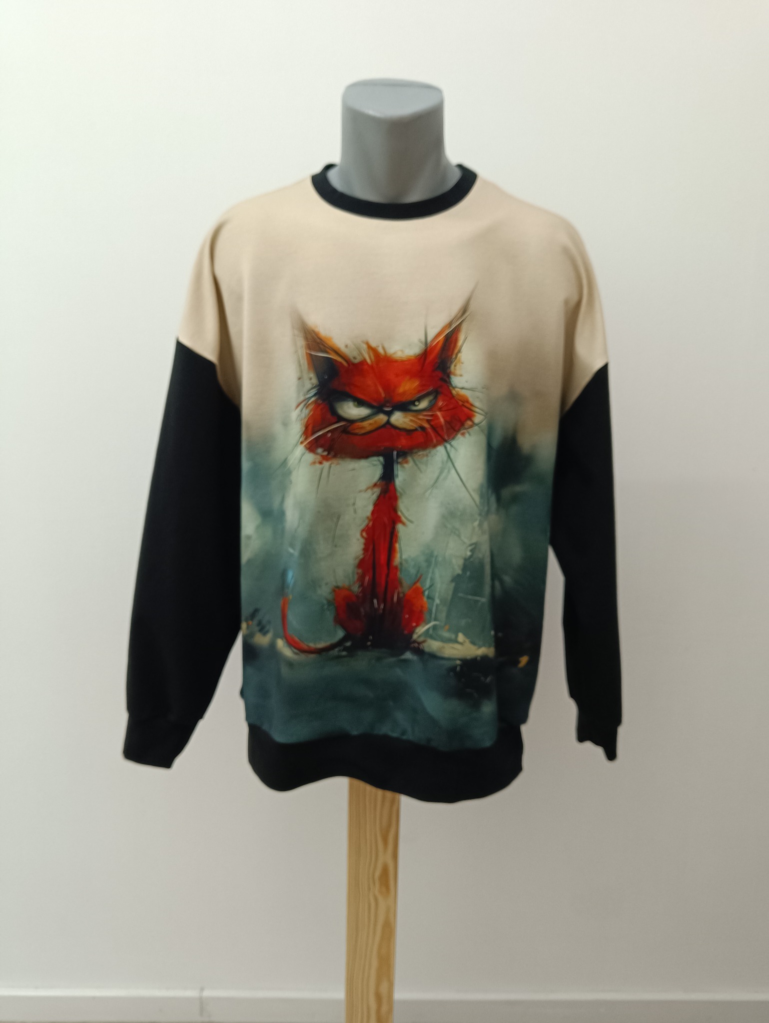 Beżowo-czarna bluza z nadrukiem wściekłego, czerwonego kota, umieszczona na manekinie na drewnianym stojaku, na tle białej ściany. Wyraźny nadruk.