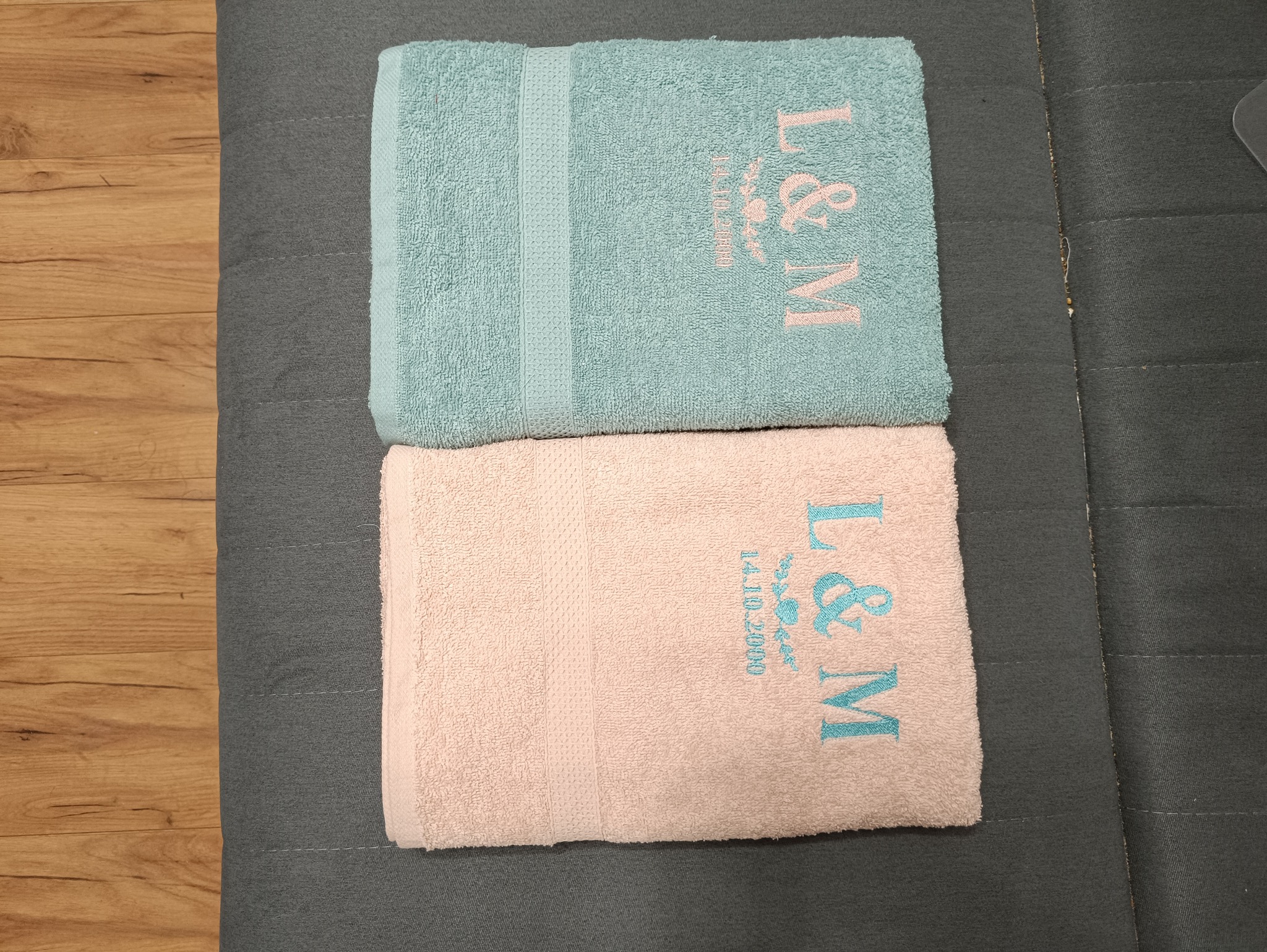 Dwa złożone ręczniki z haftem inicjałów 'L&M' i datą, leżące na szarym materiale. Ręcznik na górze jest miętowy, a na dole różowy. Widoczny fragment drewnianej podłogi.