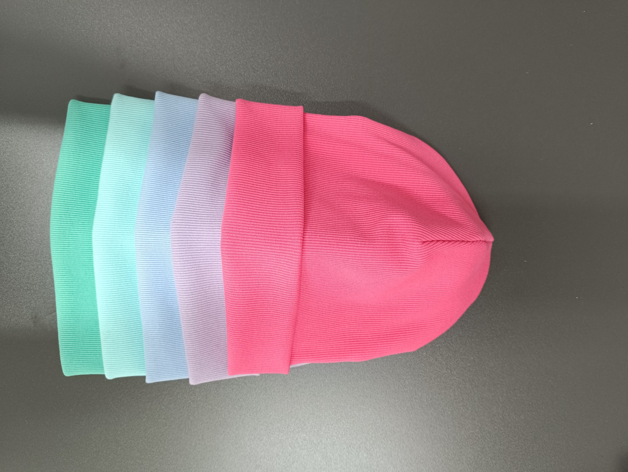 Rząd pięciu kolorowych czapek z dzianiny prążkowanej, od zieleni po róż, ułożonych na gładkim, szarym tle, prezentujących gotowe wyroby.