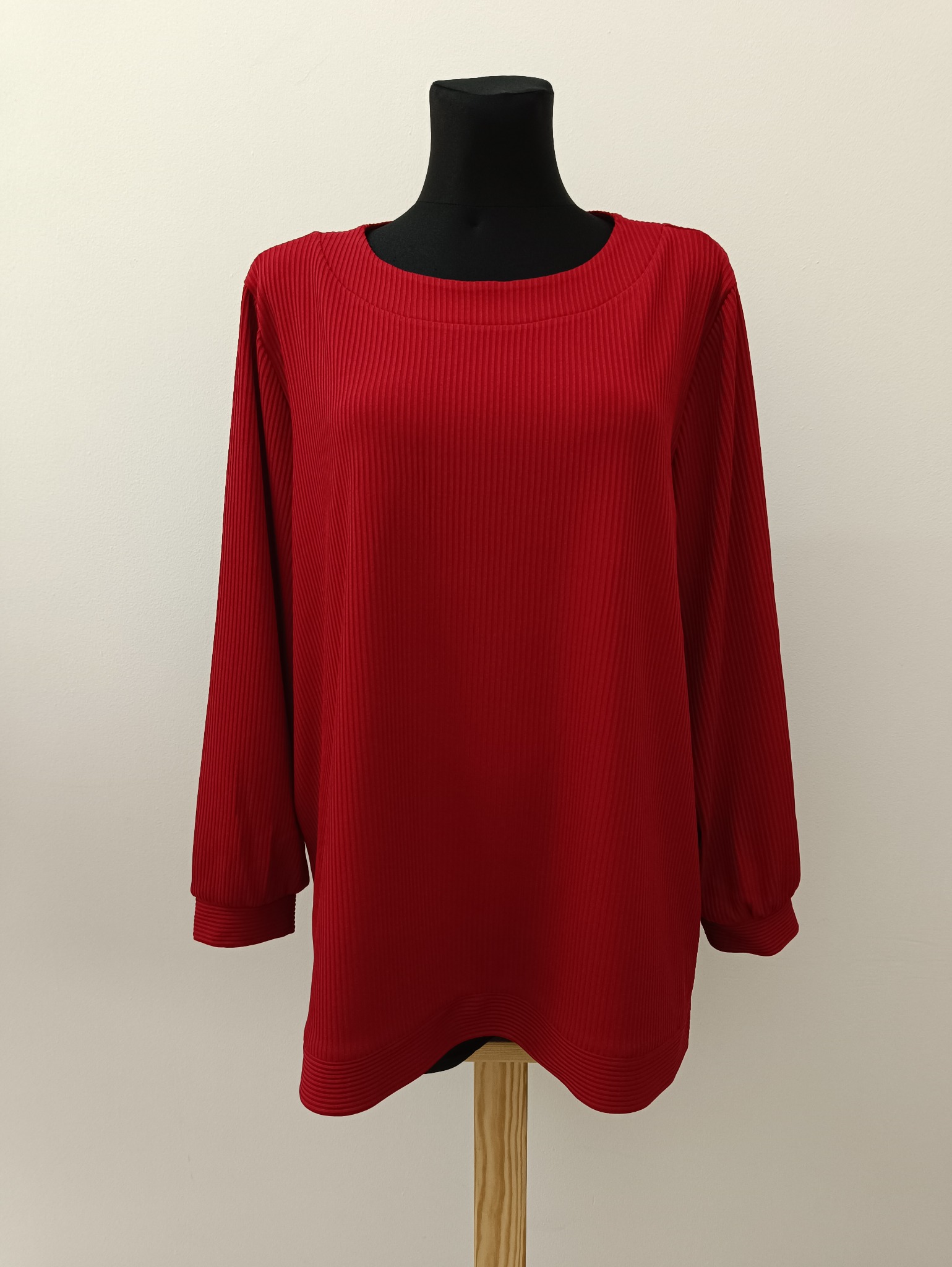 Czerwona bluzka z prążkowanej dzianiny na czarnym manekinie krawieckim. Długie rękawy z mankietami, okrągły dekolt. Neutralne tło, oświetlenie dzienne.
