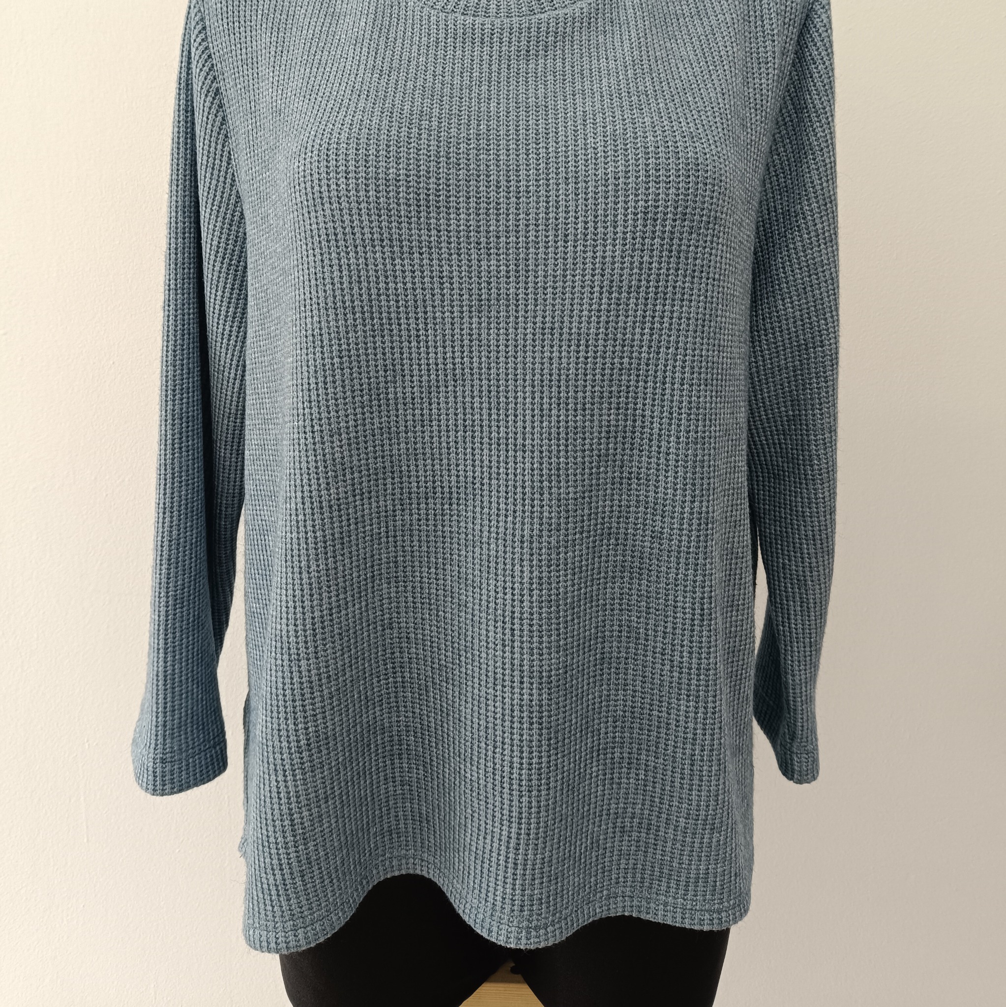 Niebieska, dzianinowa bluzka z długim rękawem na manekinie. Widoczna faktura materiału i delikatne rozcięcie po bokach. Neutralne tło.